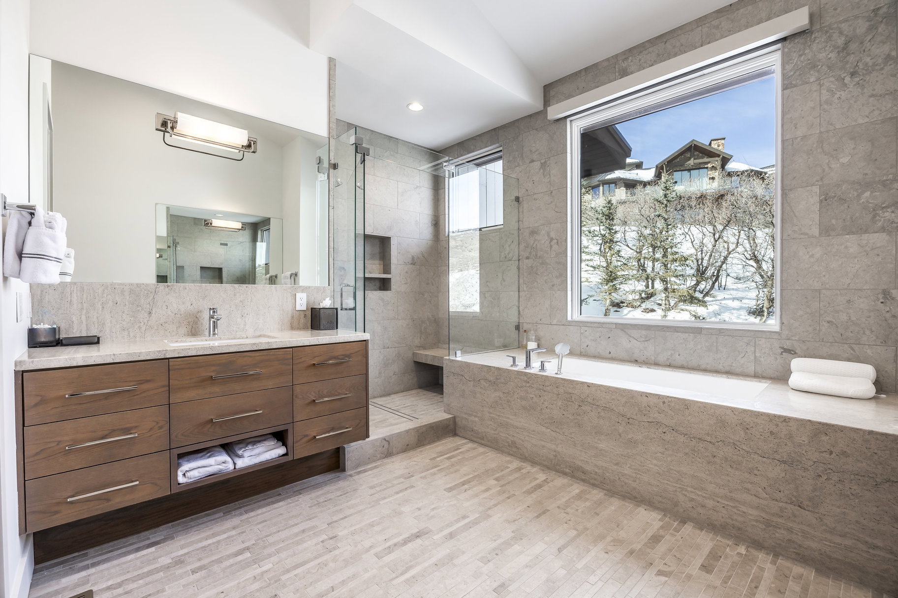 Spacious bathroom featuring a built-in soaking tub, a large picture window, a floating wood-finish vanity with an undermount sink, a glass-enclosed shower, and stone tile flooring
