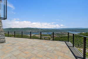 Expansive outdoor patio featuring paver flooring, modern horizontal railing, a stone-clad column, and distant water scenery