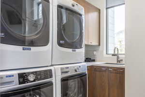 Laundry area featuring stacked appliances, a sink with a gooseneck faucet, light-toned countertop, and wood-finish cabinetry