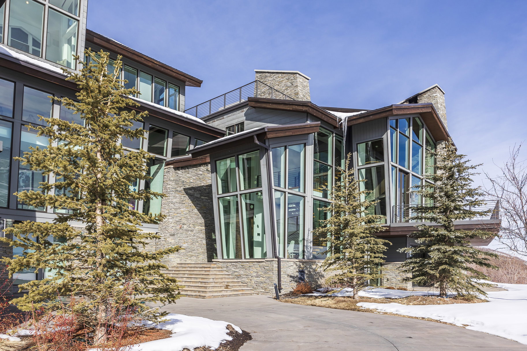 Modern architectural design featuring extensive window walls, multi-level stone facade, stone steps, and a flat roofline with a railing