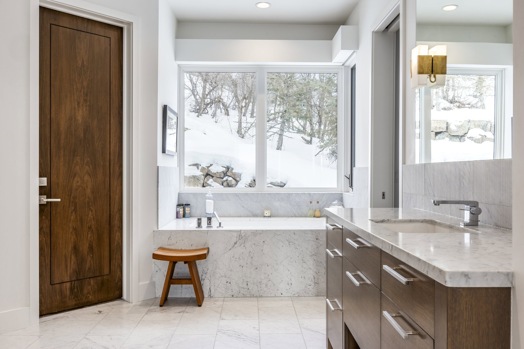 Bathroom featuring a built-in soaking tub with a tiled surround, a large window, a wood-finish vanity with a light-toned countertop, a rectangular undermount sink, and light-toned tile flooring