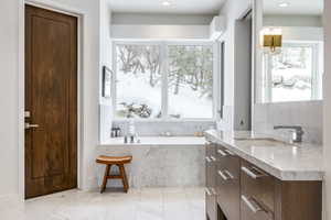 Bathroom featuring a built-in soaking tub with a tiled surround, a large window, a wood-finish vanity with a light-toned countertop, a rectangular undermount sink, and light-toned tile flooring