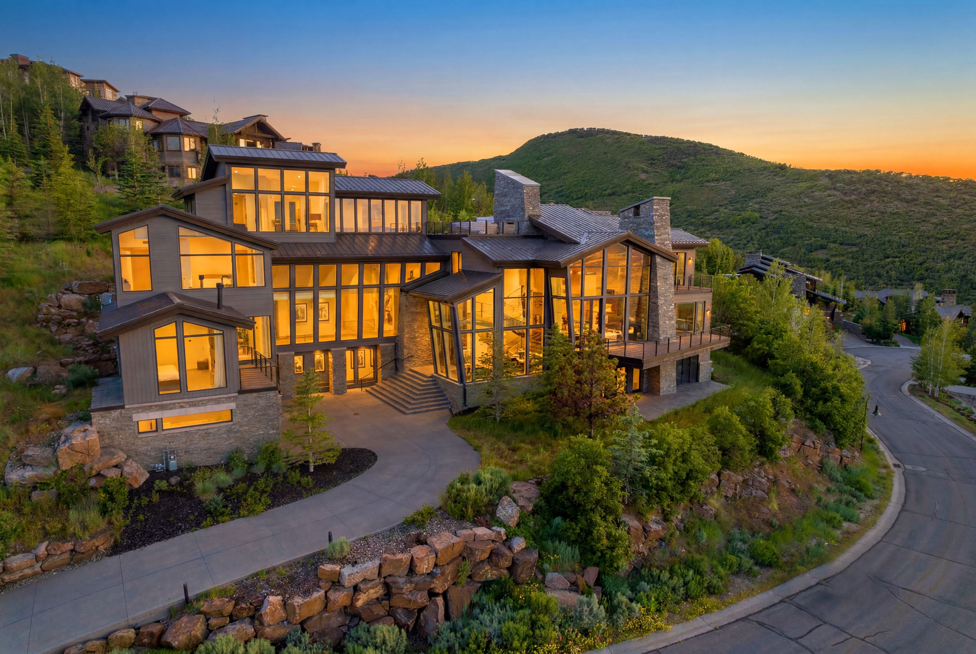 Modern architectural residence featuring extensive glazing, multiple outdoor deck spaces, stone accents, and a sloped driveway with integrated lighting