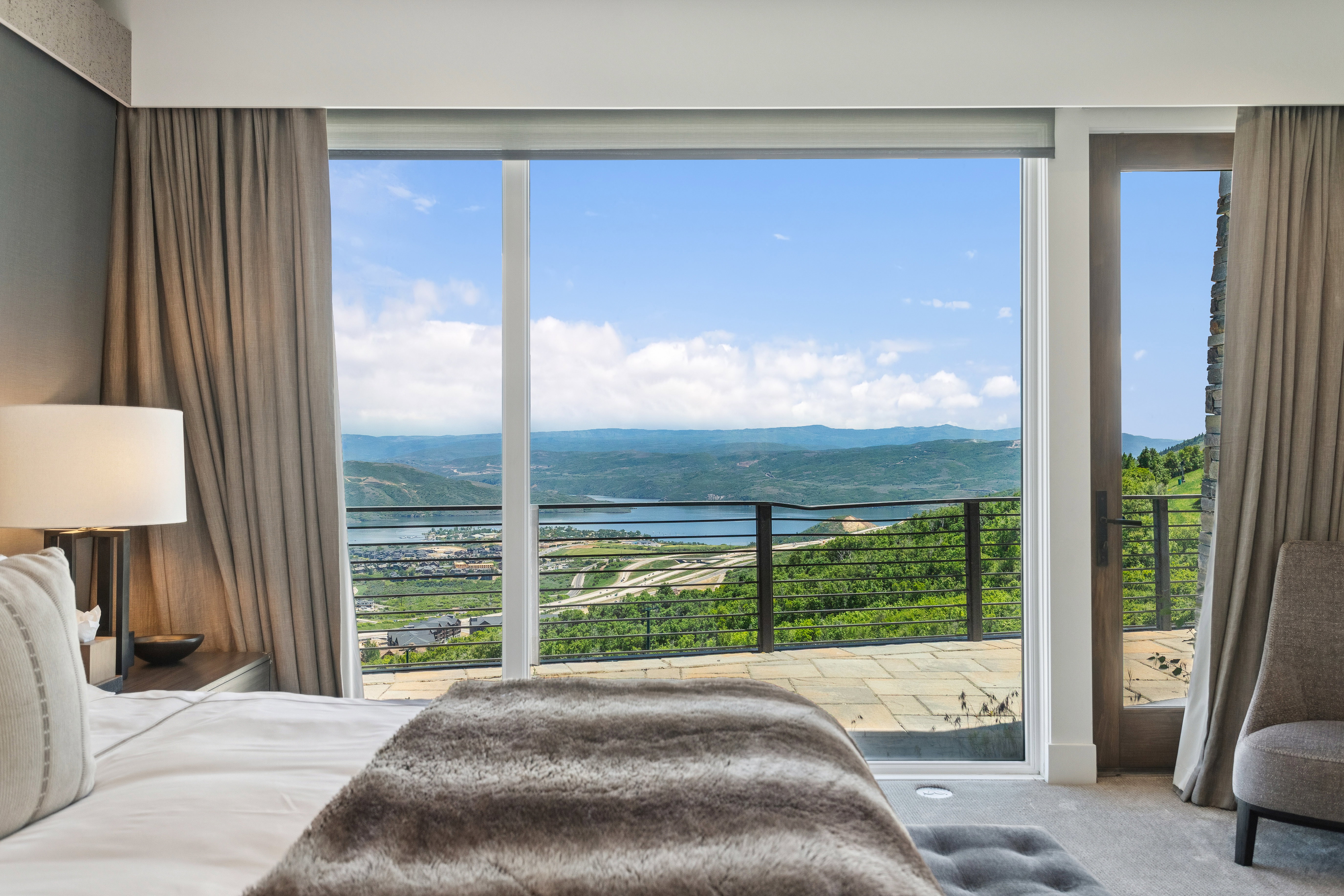 Expansive glass sliding doors open to an outdoor patio with metal railings, showcasing scenic mountain and lake vistas