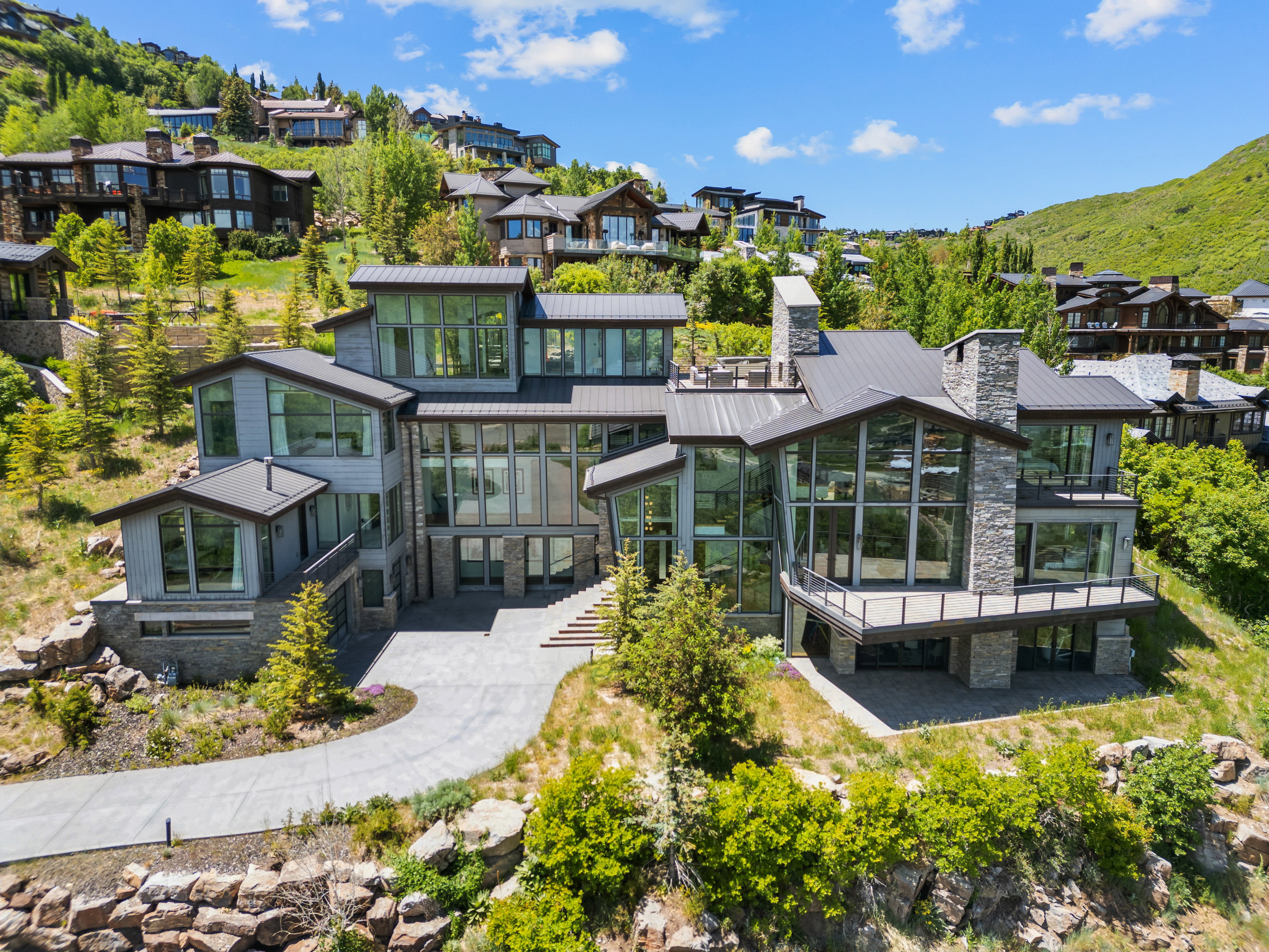Expansive modern residence featuring multiple levels, extensive glazing, a stone chimney, a large outdoor deck, and a paved driveway