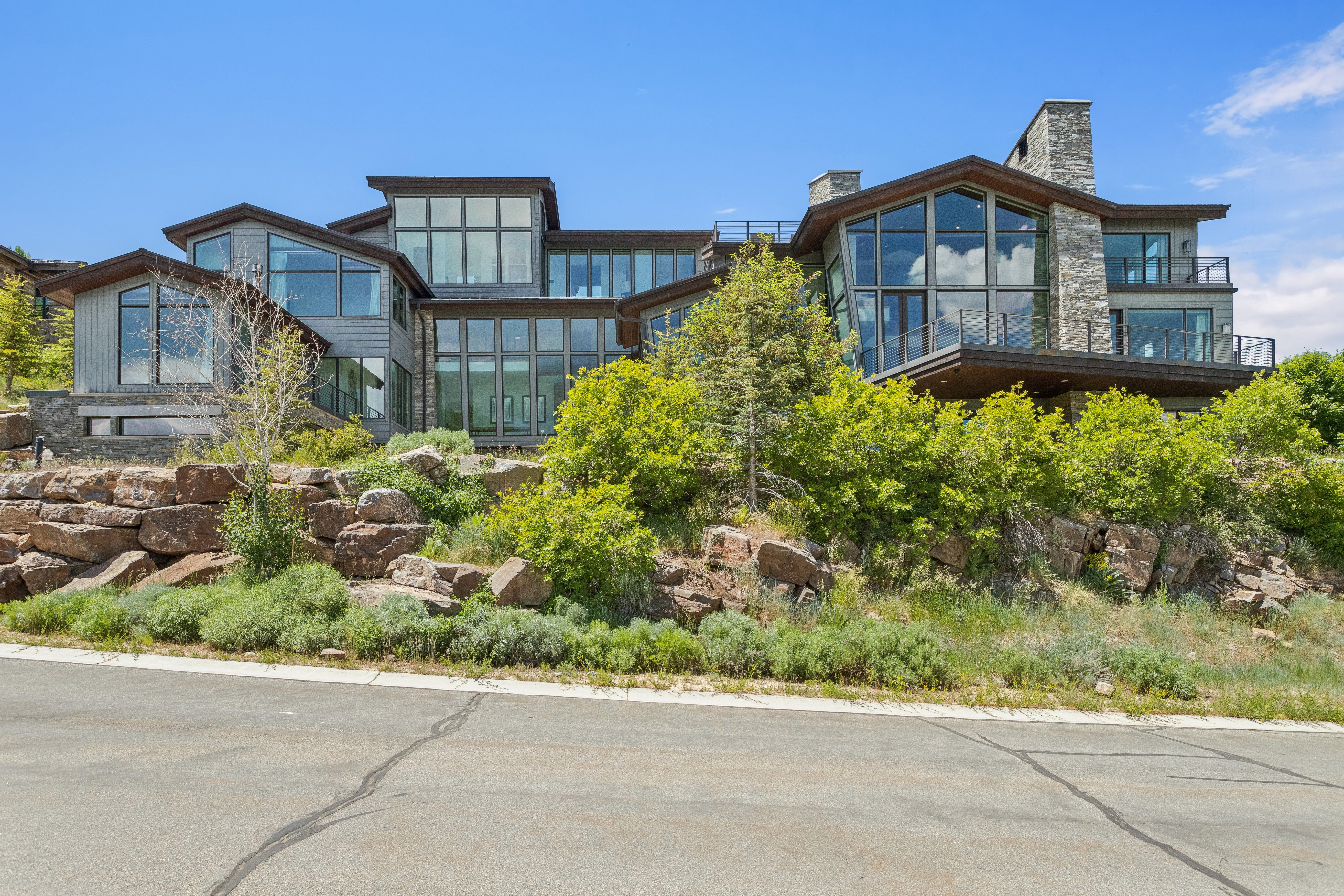 Modern multi-level residence featuring extensive floor-to-ceiling windows, stone and horizontal siding exterior finishes, multiple balconies with glass railings, and prominent stone chimneys