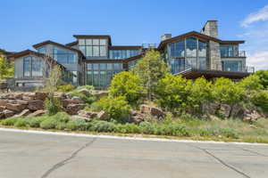 Modern multi-level residence featuring extensive floor-to-ceiling windows, stone and horizontal siding exterior finishes, multiple balconies with glass railings, and prominent stone chimneys