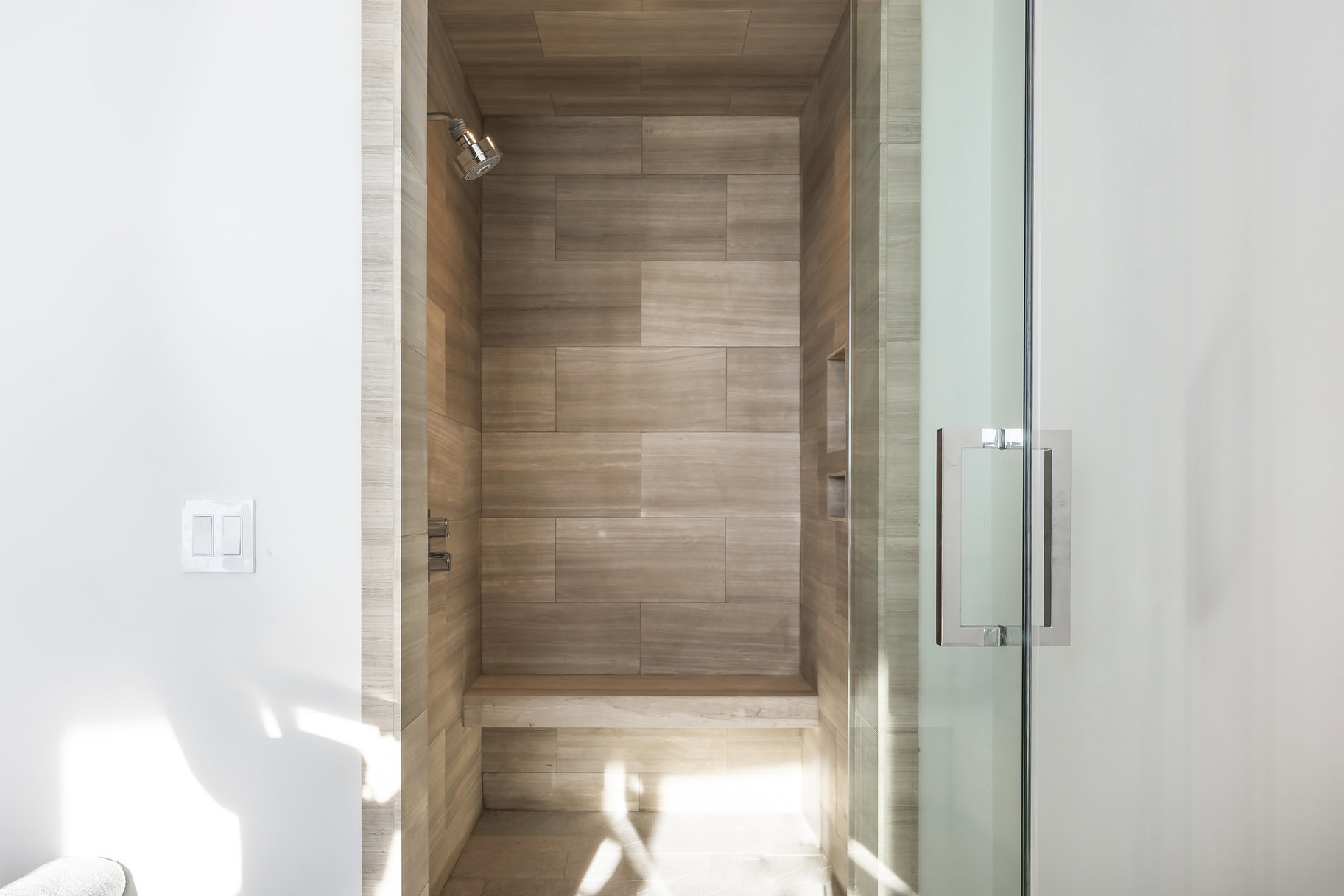 Spacious shower enclosure with floor-to-ceiling wood-look tile, a built-in bench, recessed shelving, and a glass door with a polished chrome handle