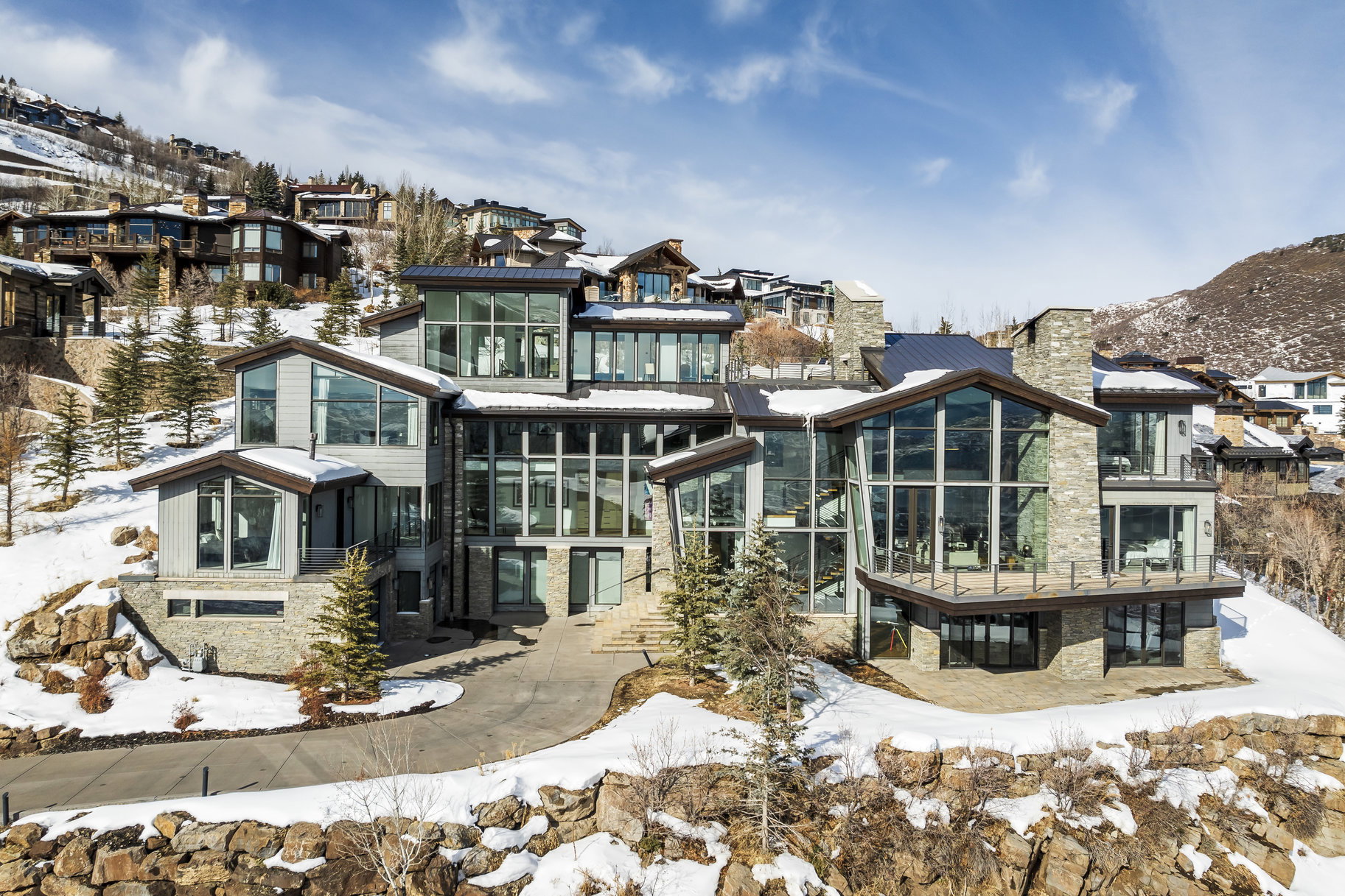 Contemporary multi-level residence featuring expansive window walls, stone veneer accents, multiple balconies with glass railings, a paved driveway, and a sloped terrain setting