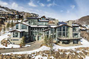 Contemporary multi-level residence featuring expansive window walls, stone veneer accents, multiple balconies with glass railings, a paved driveway, and a sloped terrain setting