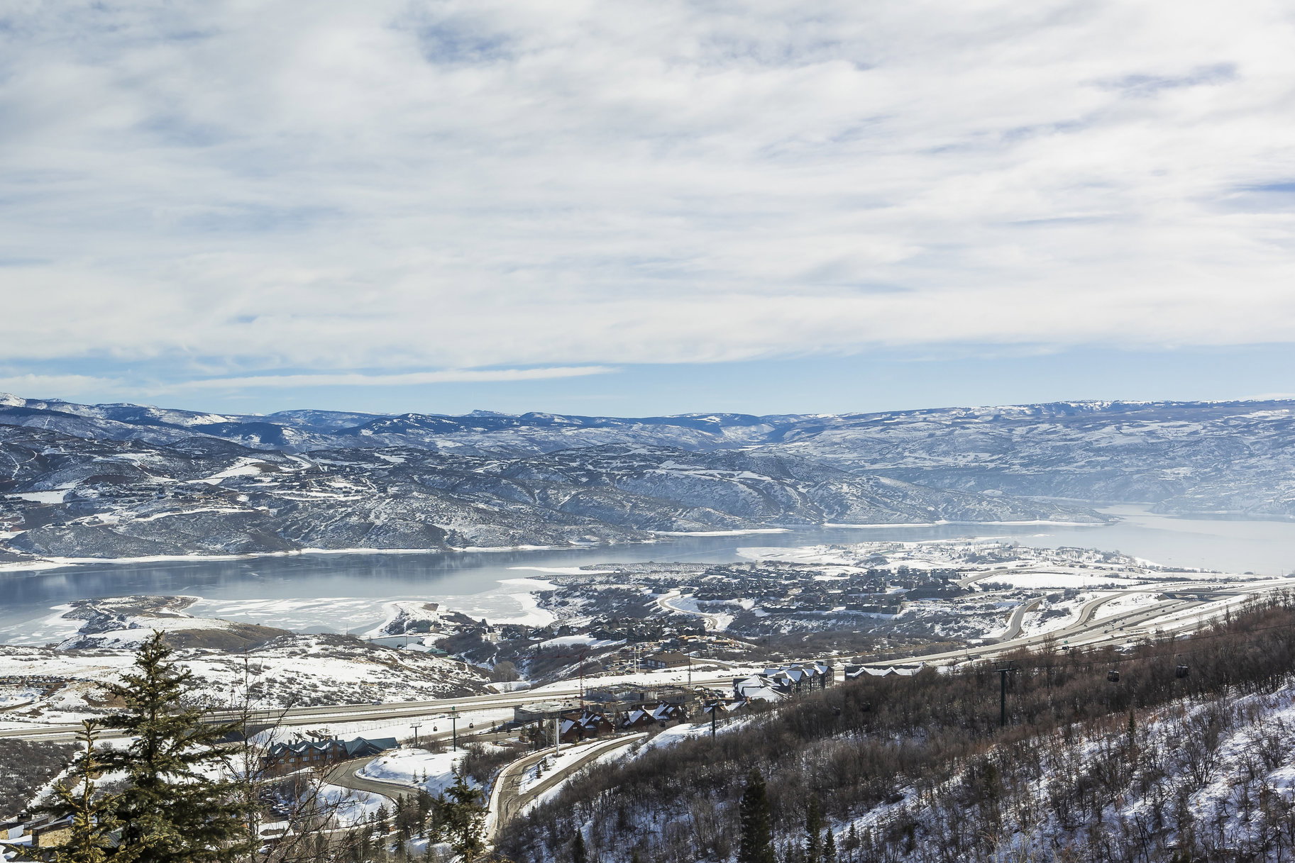 Expansive mountain landscape featuring a large body of water, snow-covered terrain, surrounding roadways, and distant structures