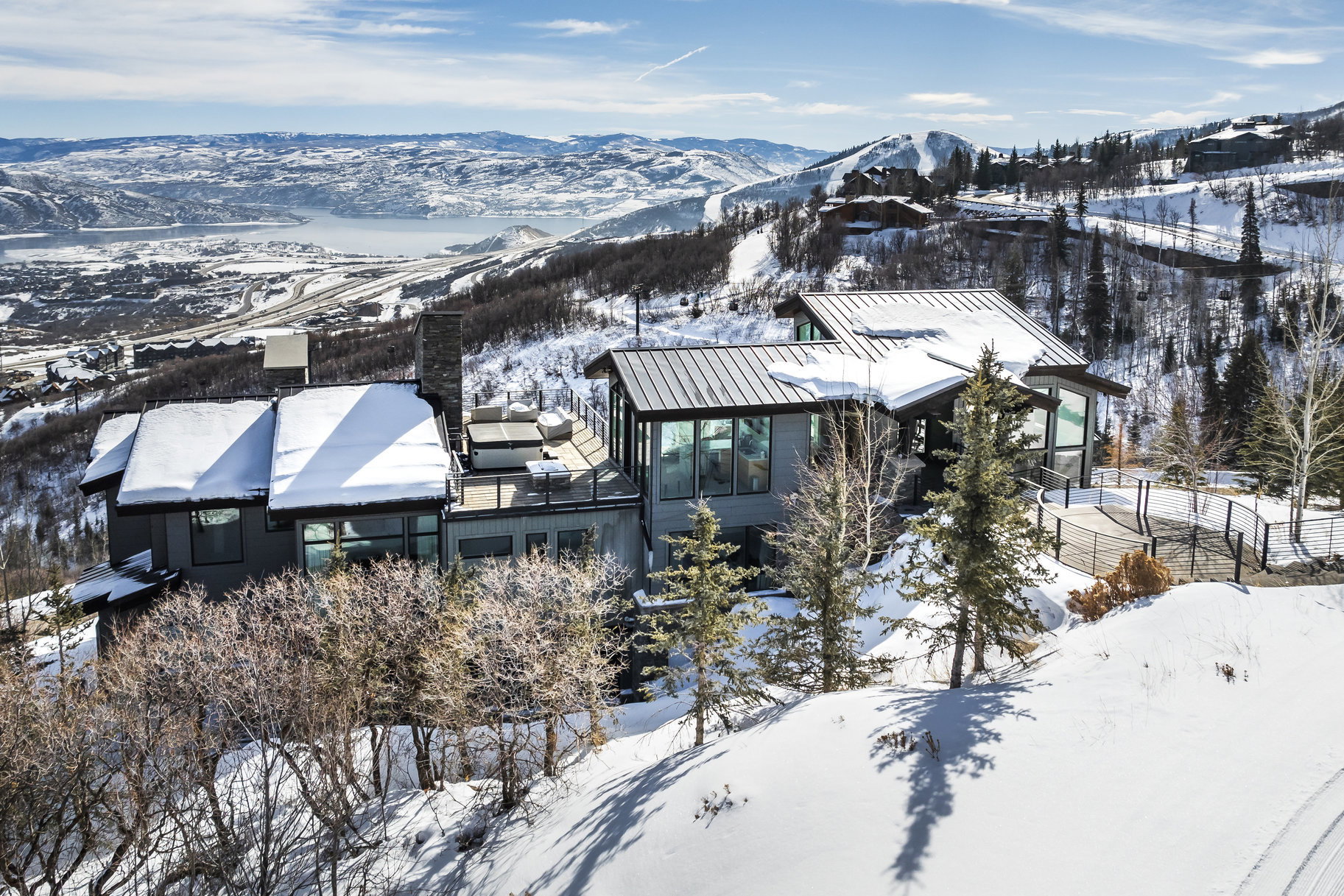 Modern mountain residence featuring an expansive deck with multiple seating areas, a prominent stone chimney, full-height windows, a metal roof, and a surrounding snow-covered landscape