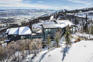 Modern mountain residence featuring an expansive deck with multiple seating areas, a prominent stone chimney, full-height windows, a metal roof, and a surrounding snow-covered landscape