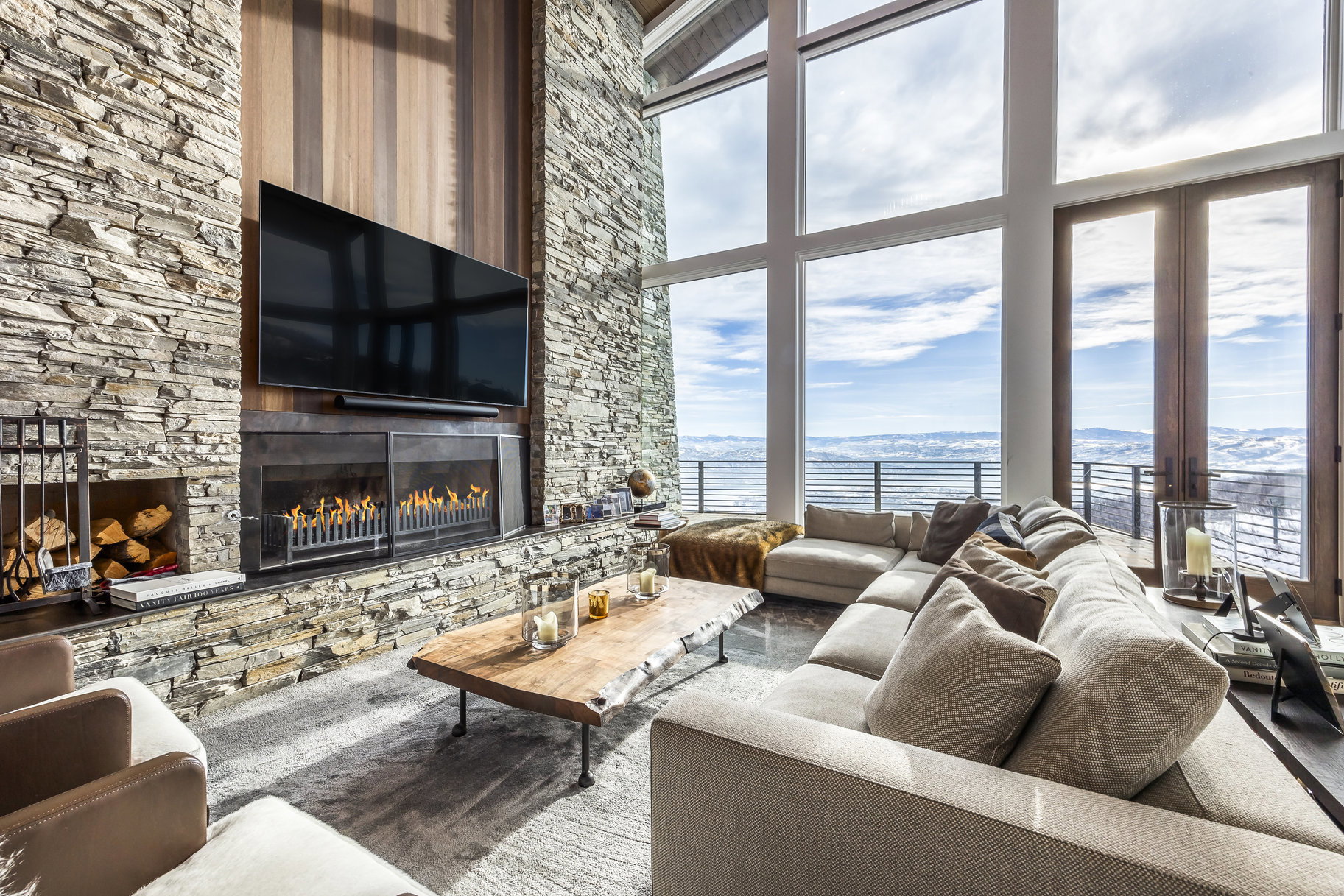 Spacious living area featuring a floor-to-ceiling stone fireplace, large format windows, high ceilings, a built-in linear fireplace, and a wood-paneled accent wall
