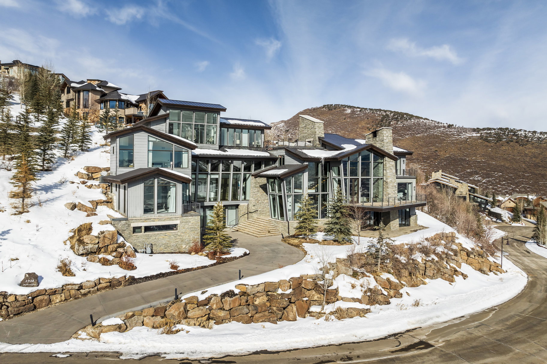 Contemporary multi-level residence featuring extensive floor-to-ceiling windows, stone and siding exterior finishes, multiple balconies, a terraced landscape, and a winding driveway