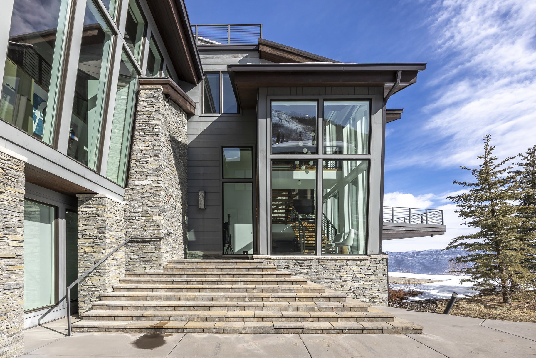 Contemporary exterior featuring stone cladding, multi-pane windows, a prominent glass facade, a large outdoor deck, and a stone staircase