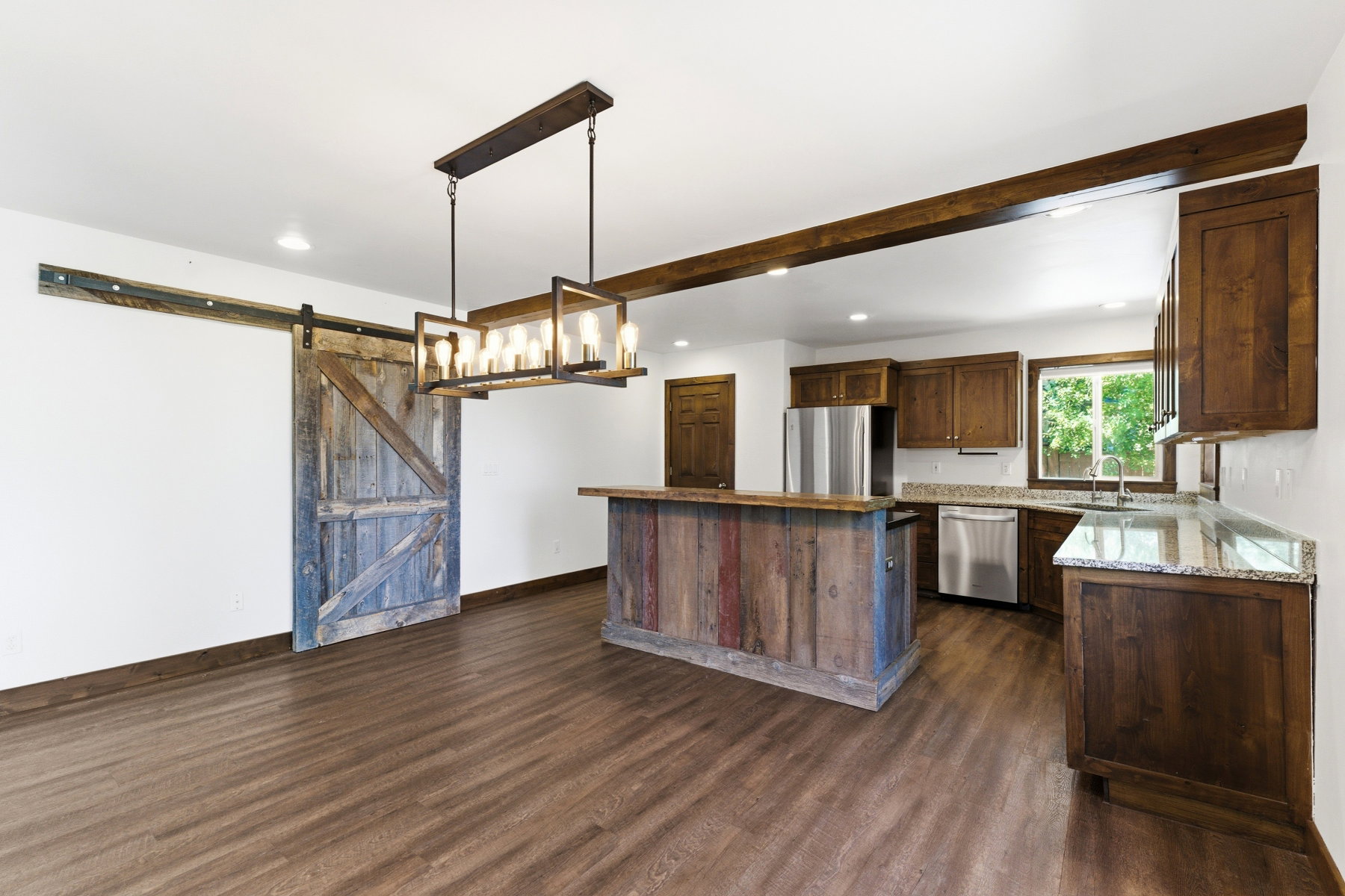 Kitchen and dining area featuring wood-finish flooring, a rustic barn door, a substantial wood-beam ceiling accent, a reclaimed wood-clad island, and stainless steel appliances
