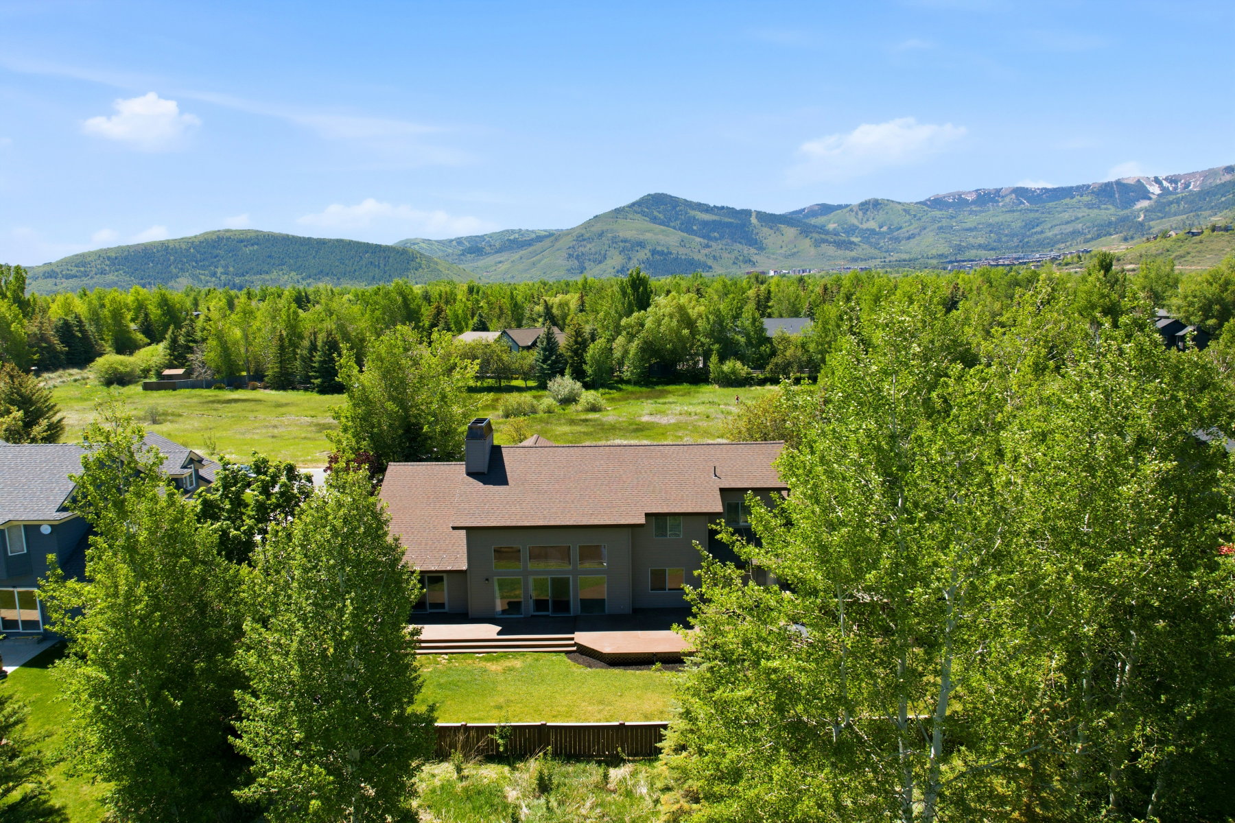 Mountain landscape featuring a residential property with a brown shingle roof, a large wooden deck, and extensive green lawn