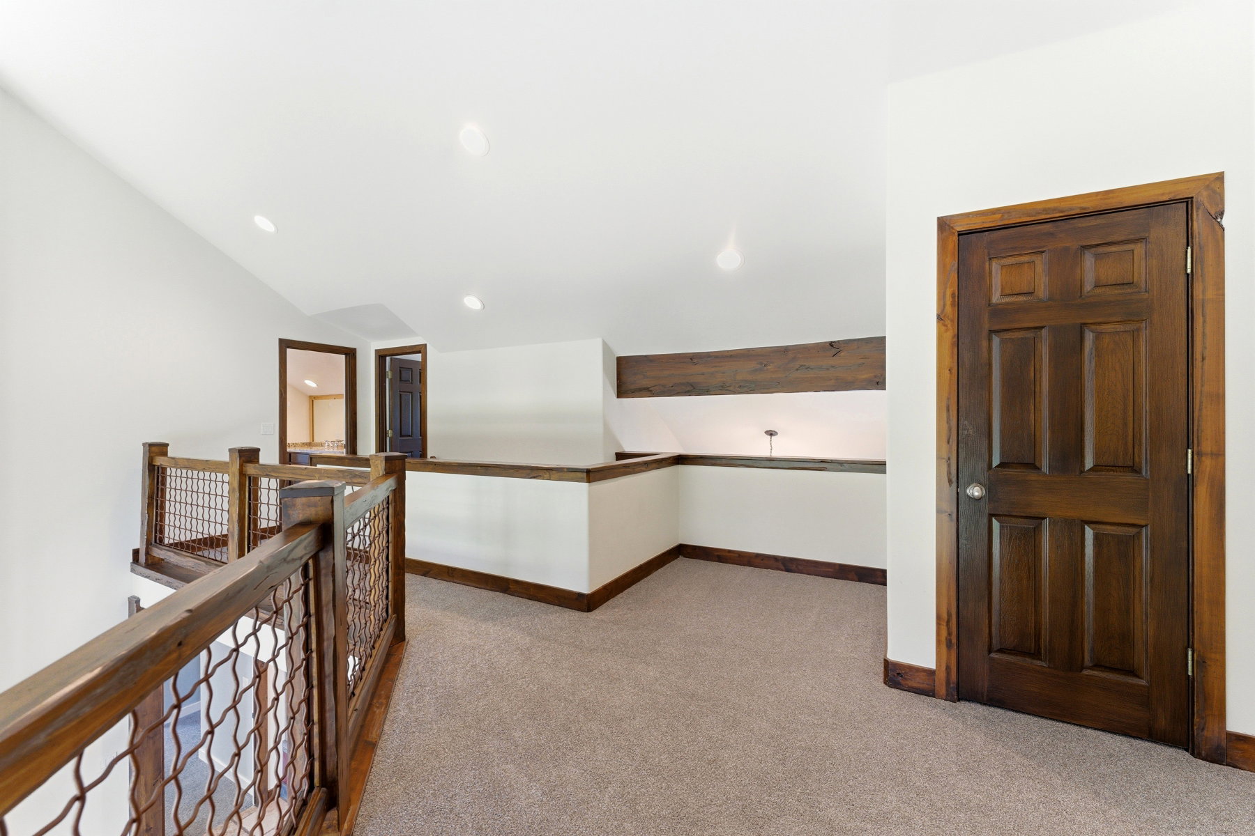 Upper-level landing featuring textured carpet flooring, wood railing with metal infill panels, recessed lighting, a wooden door, and a built-in shelf with wood trim
