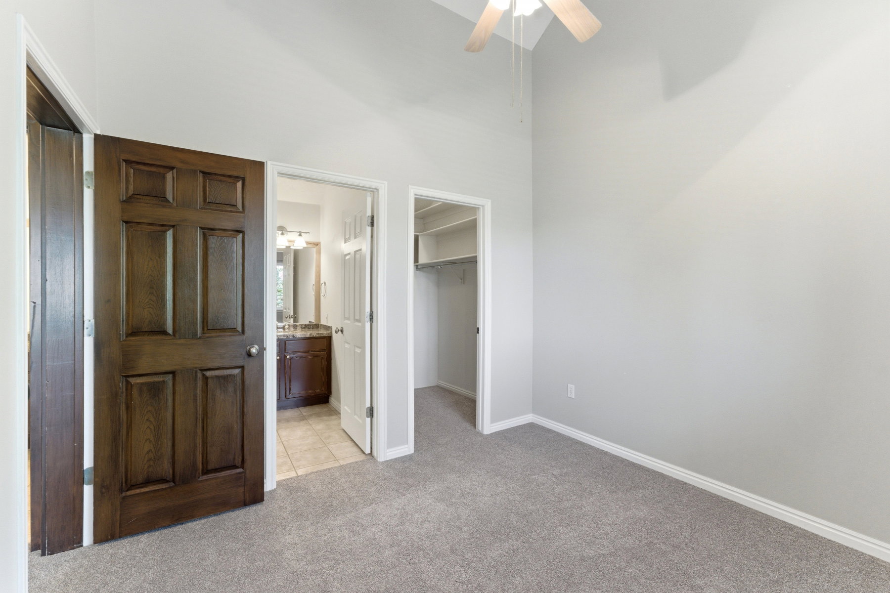 Carpeted room featuring a vaulted ceiling with a ceiling fan, a walk-in closet with built-in shelving, and a private bathroom entry with a dark wood vanity and tiled flooring