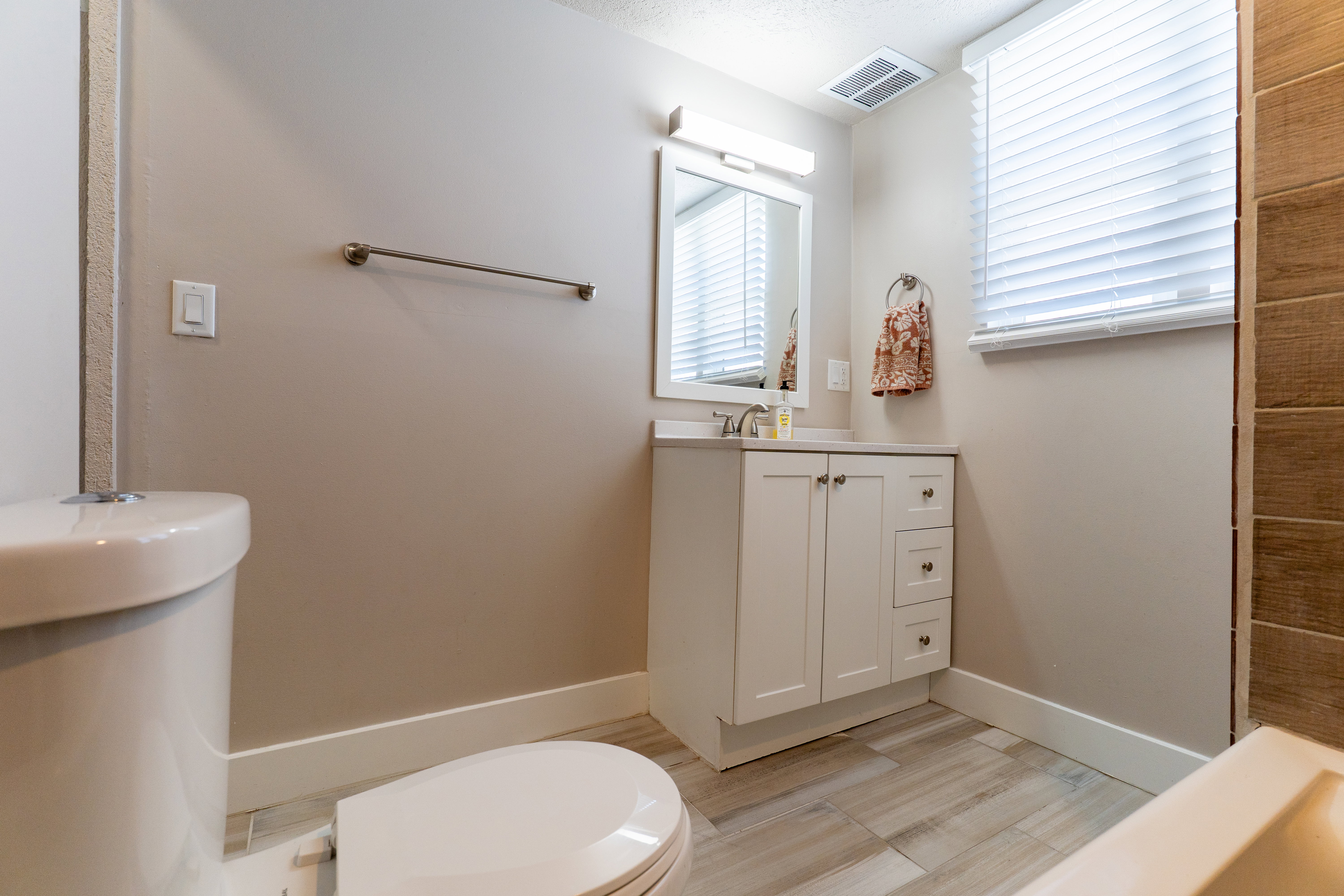 Bathroom featuring a white single vanity with integrated sink, a rectangular mirror with overhead lighting, a toilet, wood-finish flooring, and a window with blinds