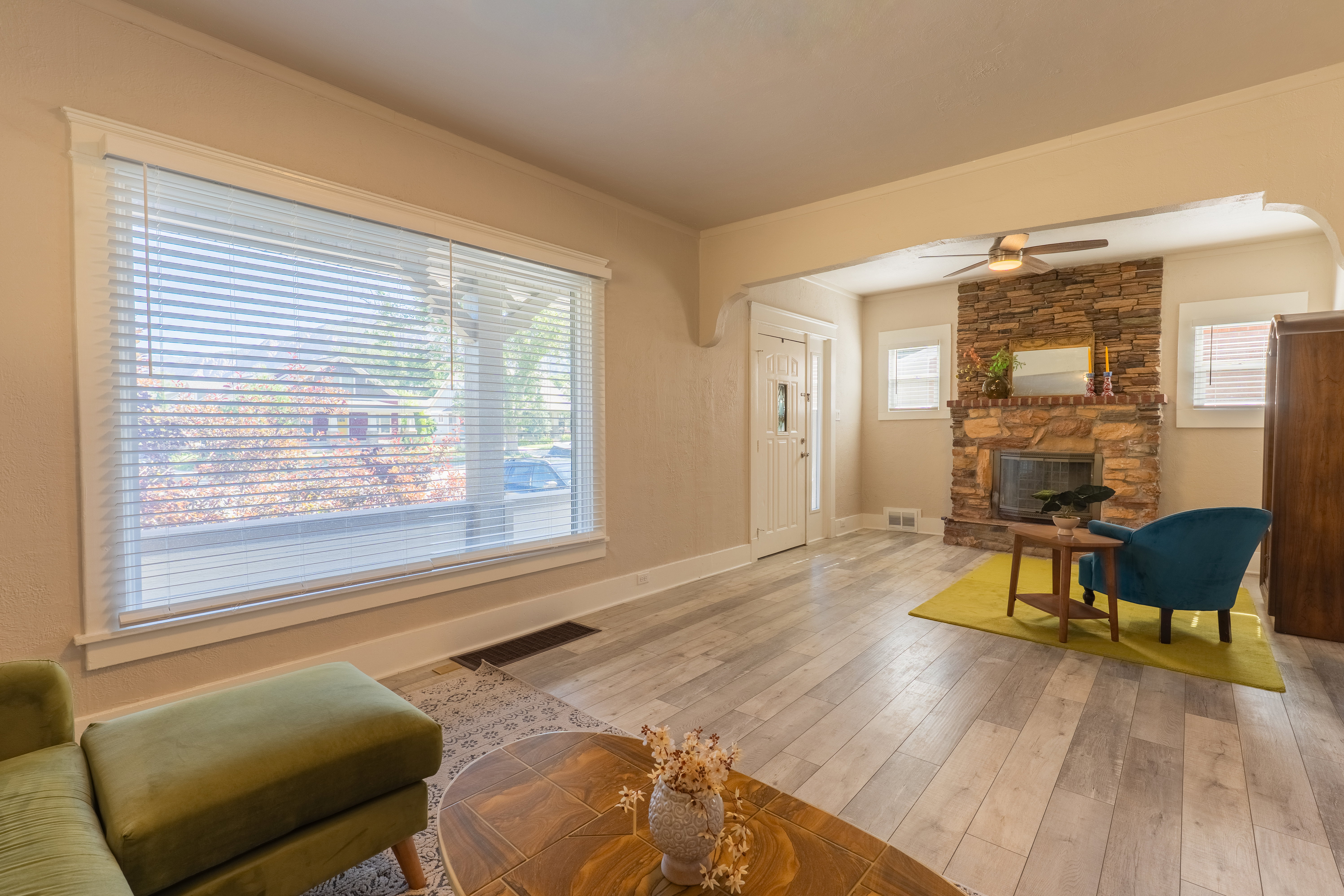 Spacious living area featuring light wood-finish flooring, a stone-faced fireplace, a ceiling fan, and large windows with blinds