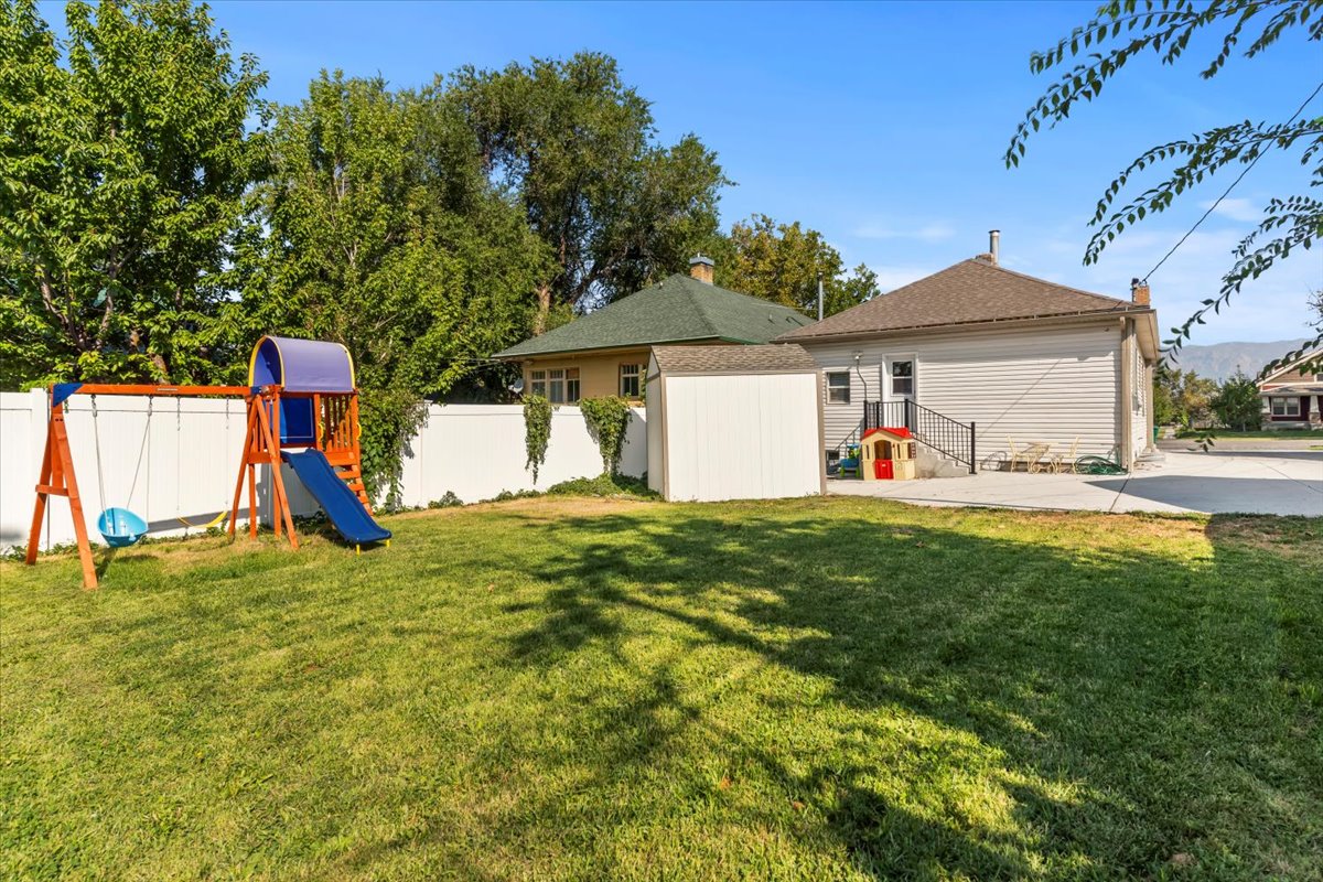 Expansive backyard featuring a lush lawn, white privacy fencing, mature trees, a concrete patio, and a dedicated storage shed