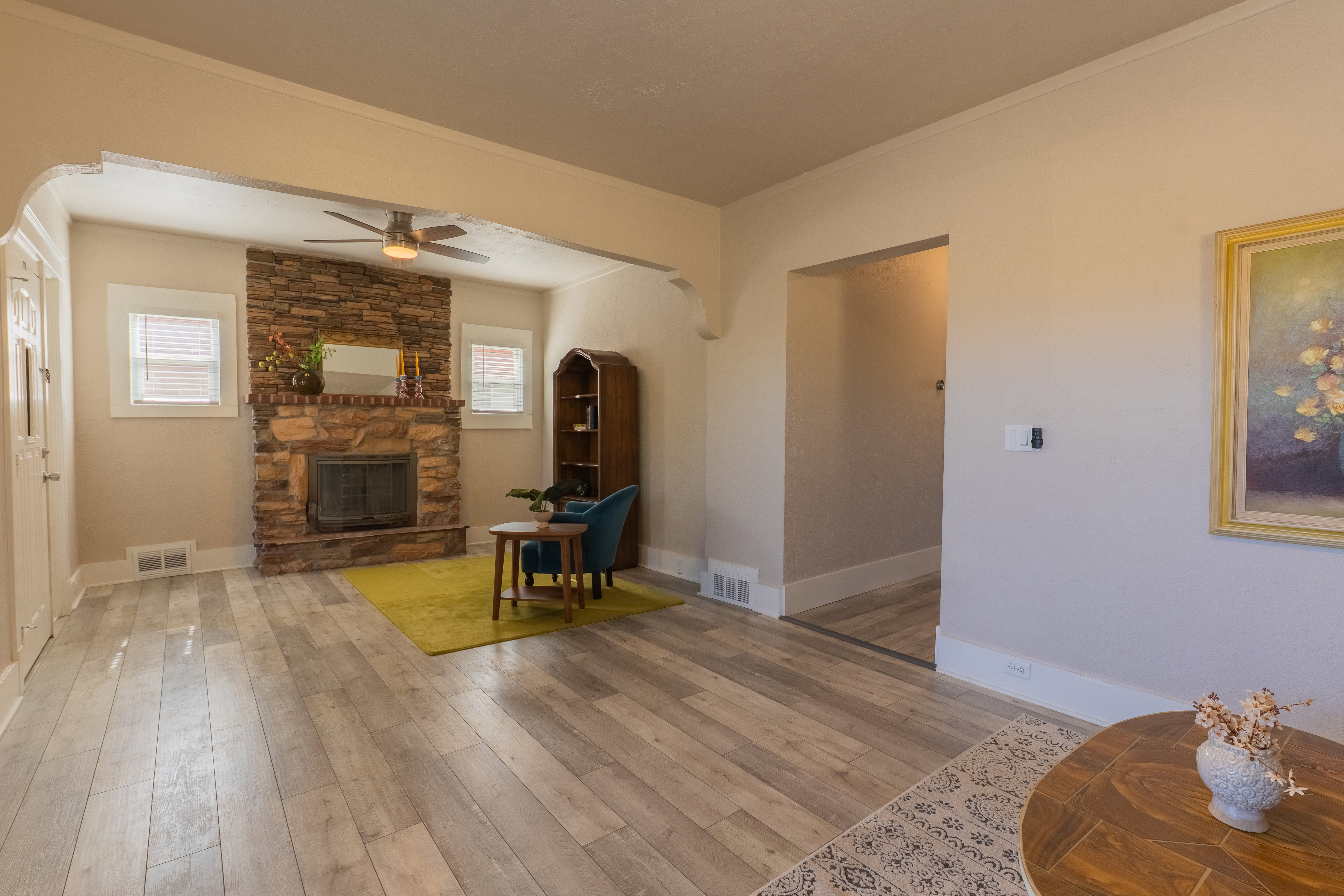 Living area featuring a stone-clad fireplace with hearth, wood-finish flooring, two large windows with blinds, a ceiling fan, and decorative arched doorways