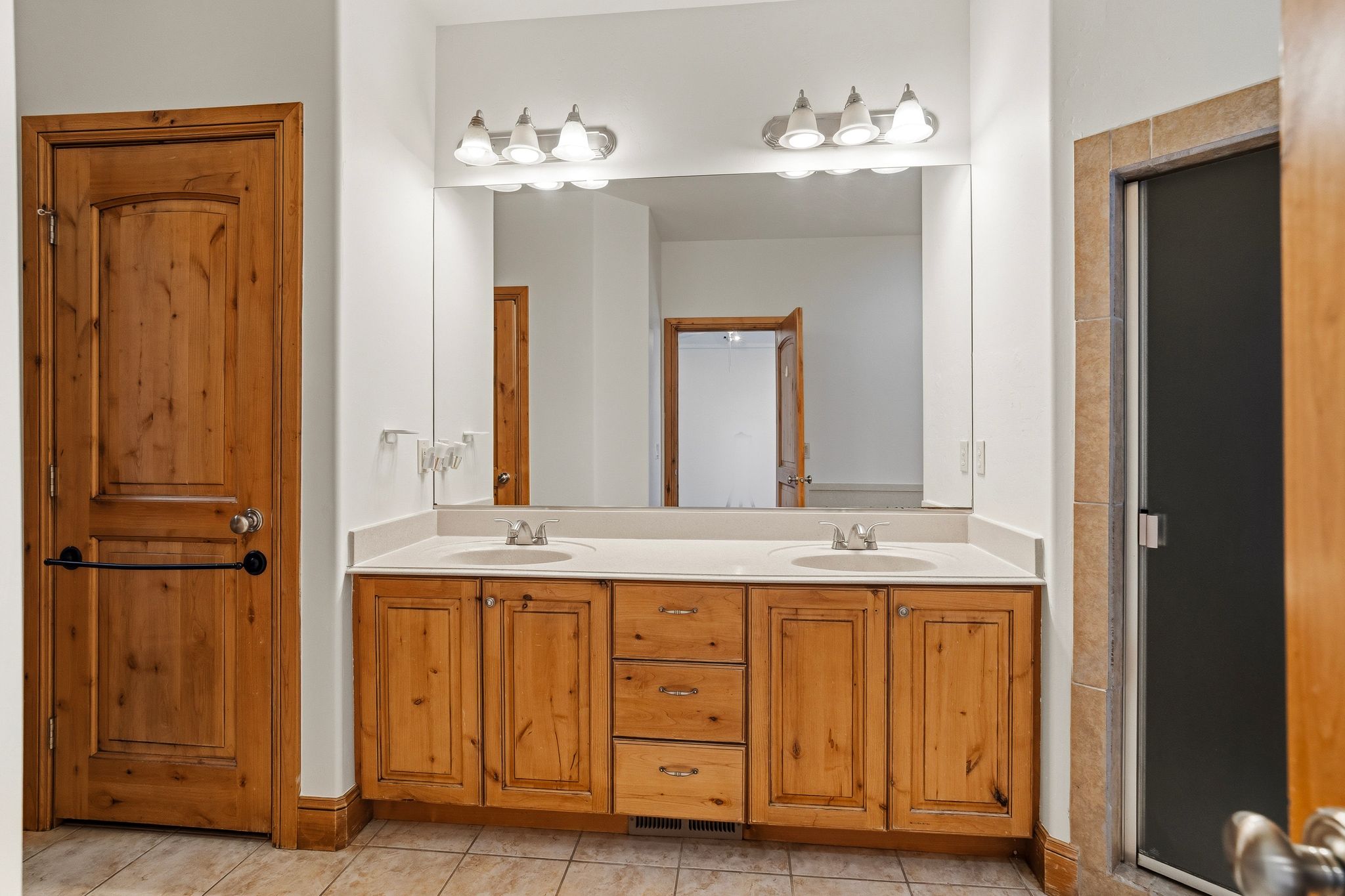 Bathroom featuring a dual vanity with light-toned wood cabinetry and a light countertop, two rectangular mirrors, dual vanity lighting, and a tiled floor