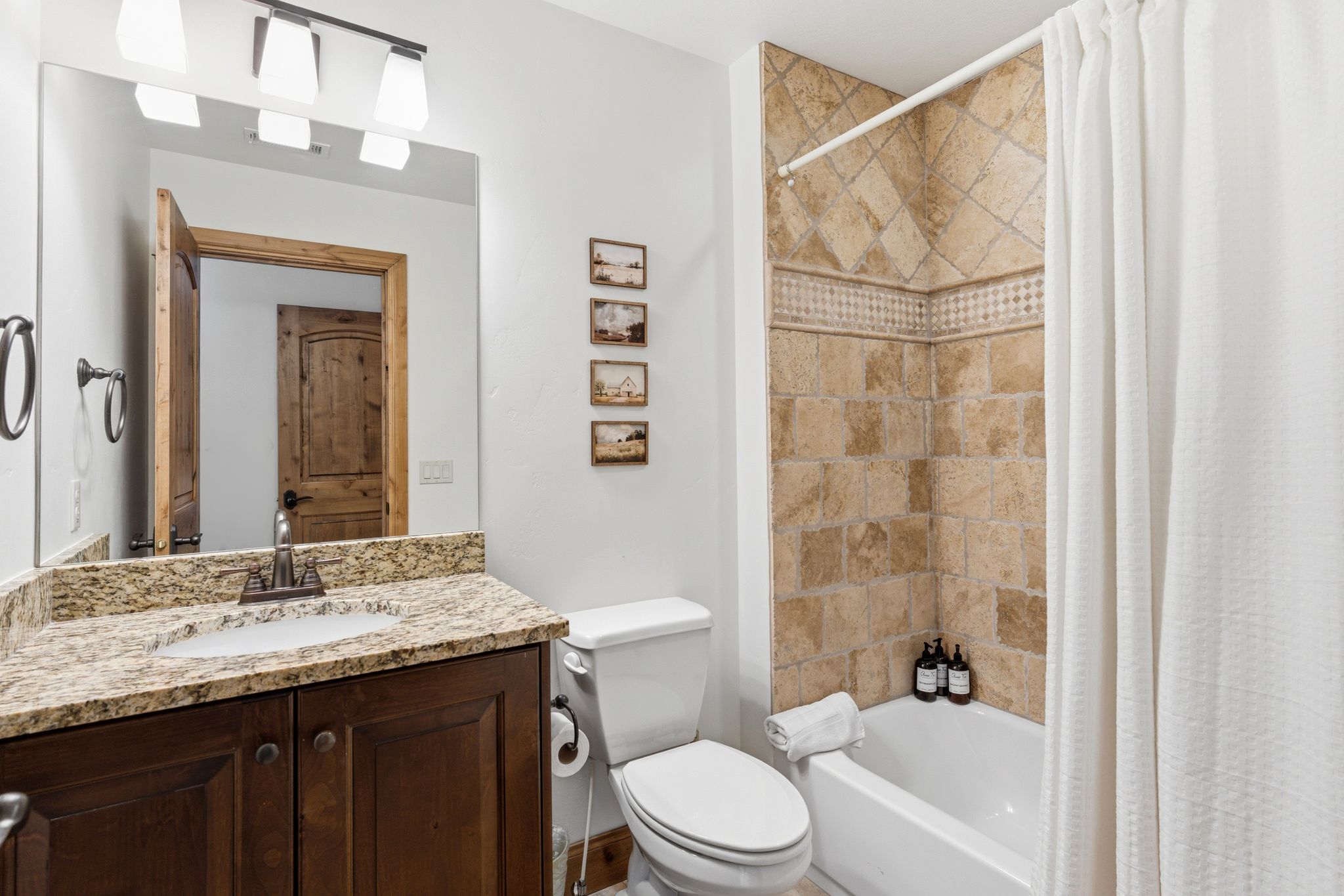 Bathroom featuring a dark wood vanity with a light-colored stone countertop, an integrated sink with a brushed nickel faucet, a large wall-mounted mirror, and a tiled shower-over-tub with a decorative mosaic accent band