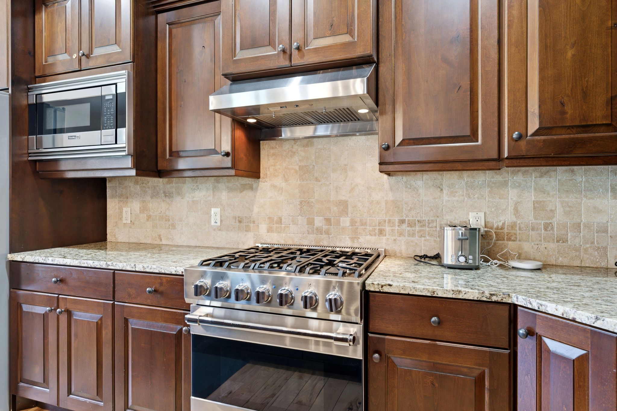 Kitchen featuring a stainless steel range with a coordinating range hood, full-height tiled backsplash with mosaic accents, granite countertops, and rich wood cabinetry