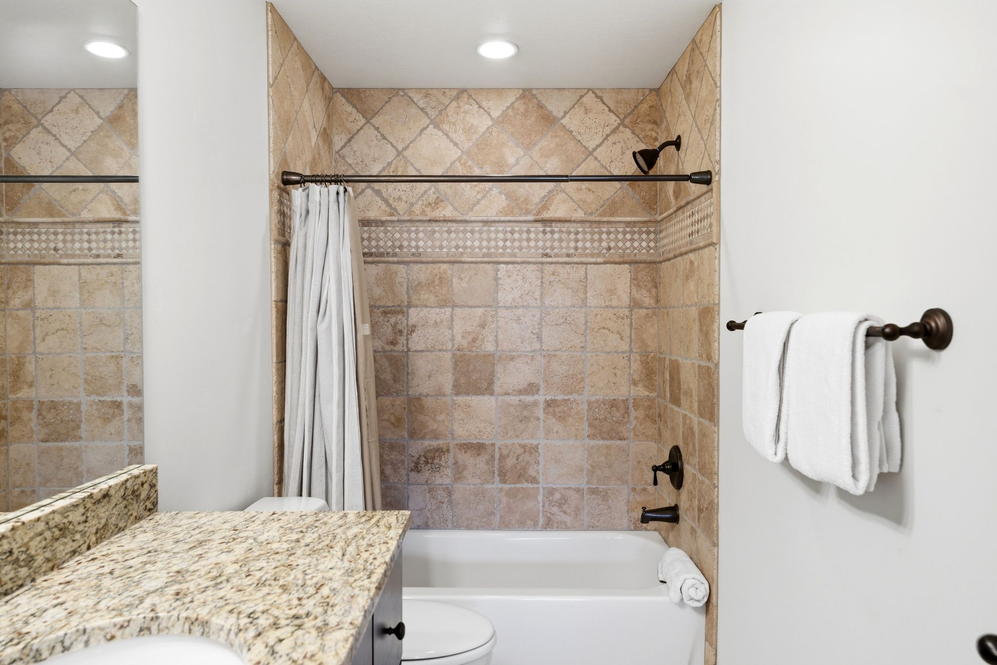 Bathroom featuring a tub/shower combination with tile surround and decorative accent band, bronze-finish fixtures, and a granite-finish vanity with an undermount sink