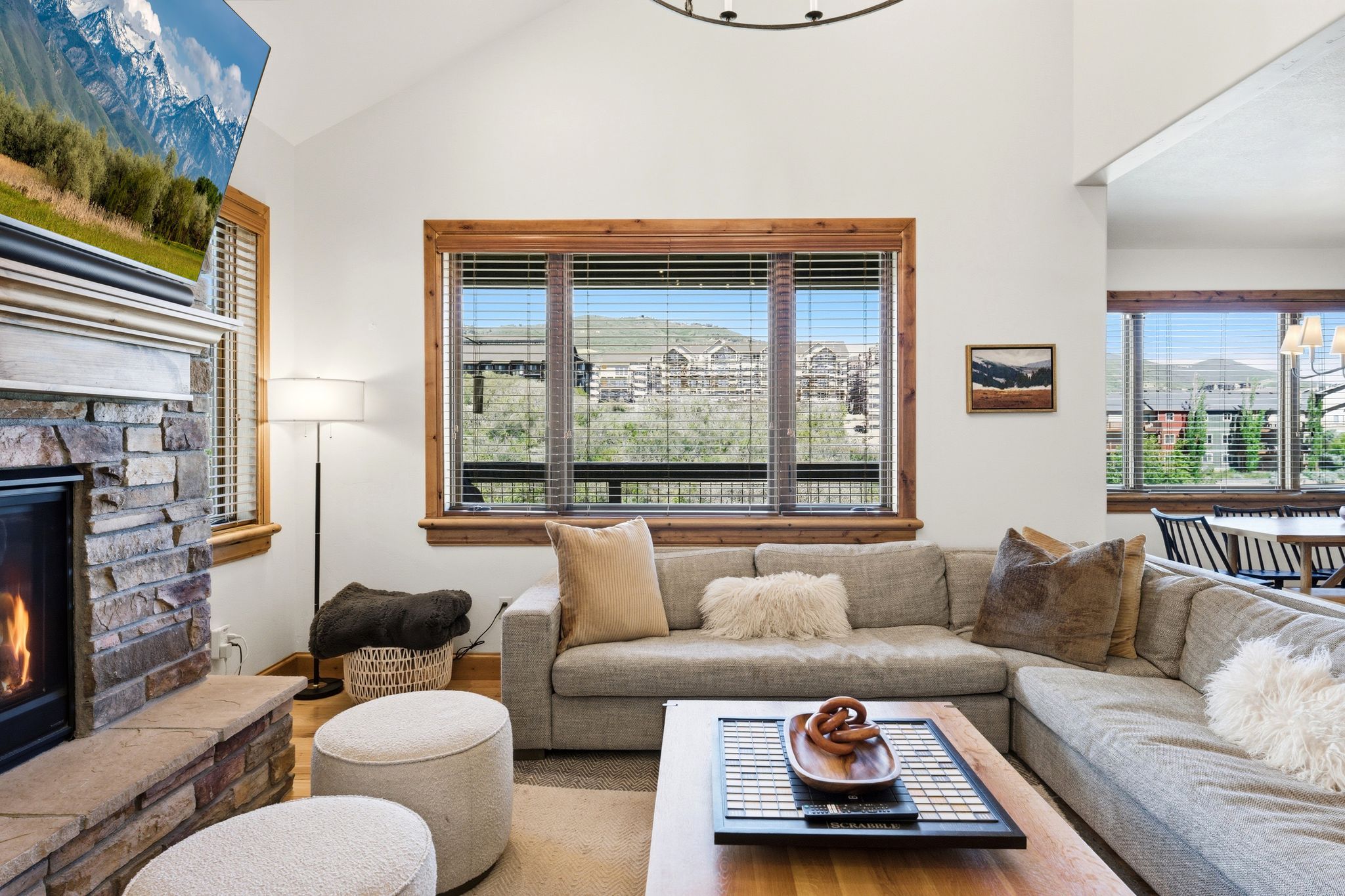 Vaulted ceilings complement an open living space featuring a stone-clad fireplace with a hearth, natural wood window trim, and multiple large windows with slat blinds
