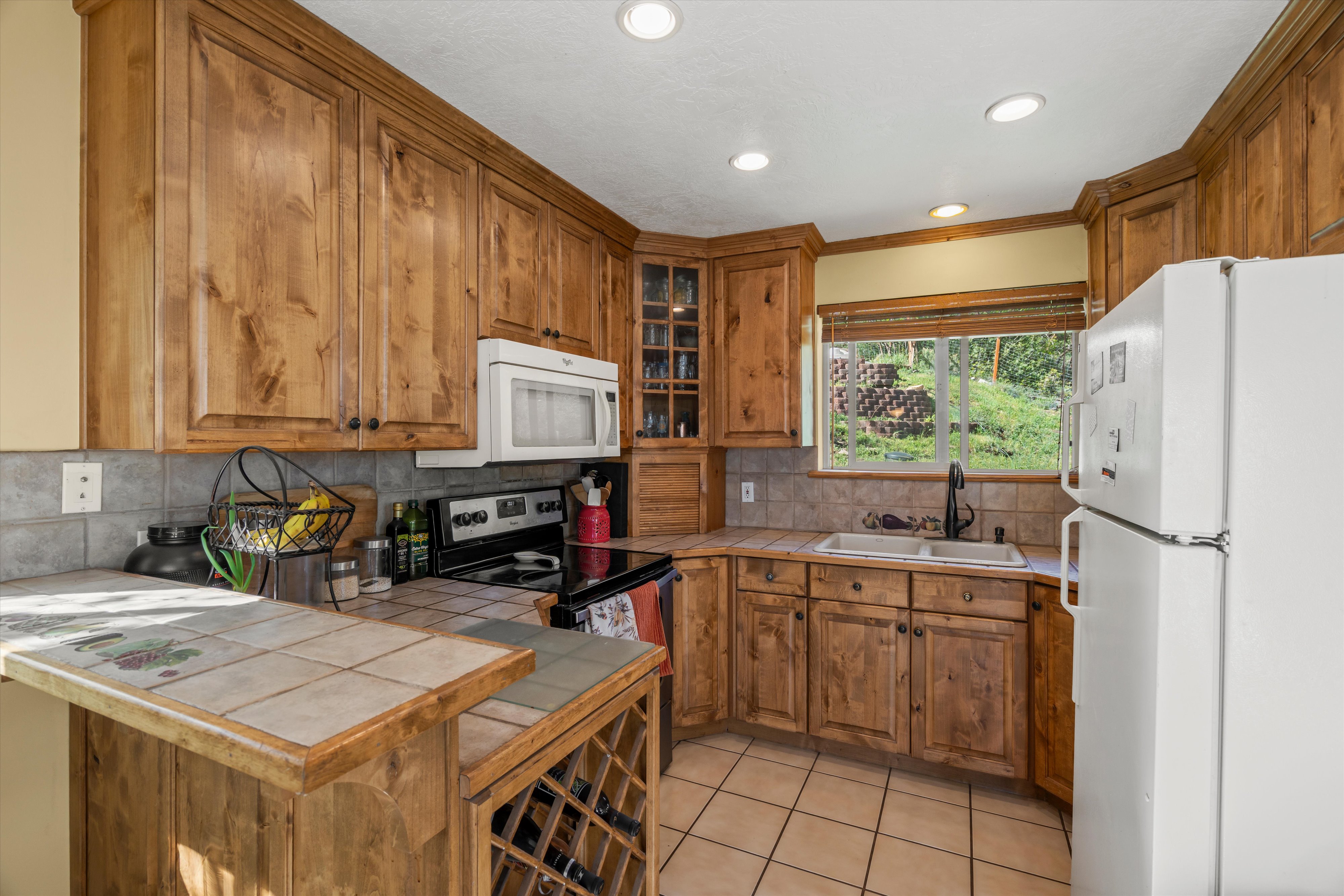 Kitchen featuring wood cabinetry, tile countertops, tile backsplash, and a window overlooking the garden and natural landscape