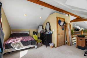 Carpeted room featuring a white headboard, recessed lighting, exposed ceiling beams, a natural wood paneled door, and a wooden railing