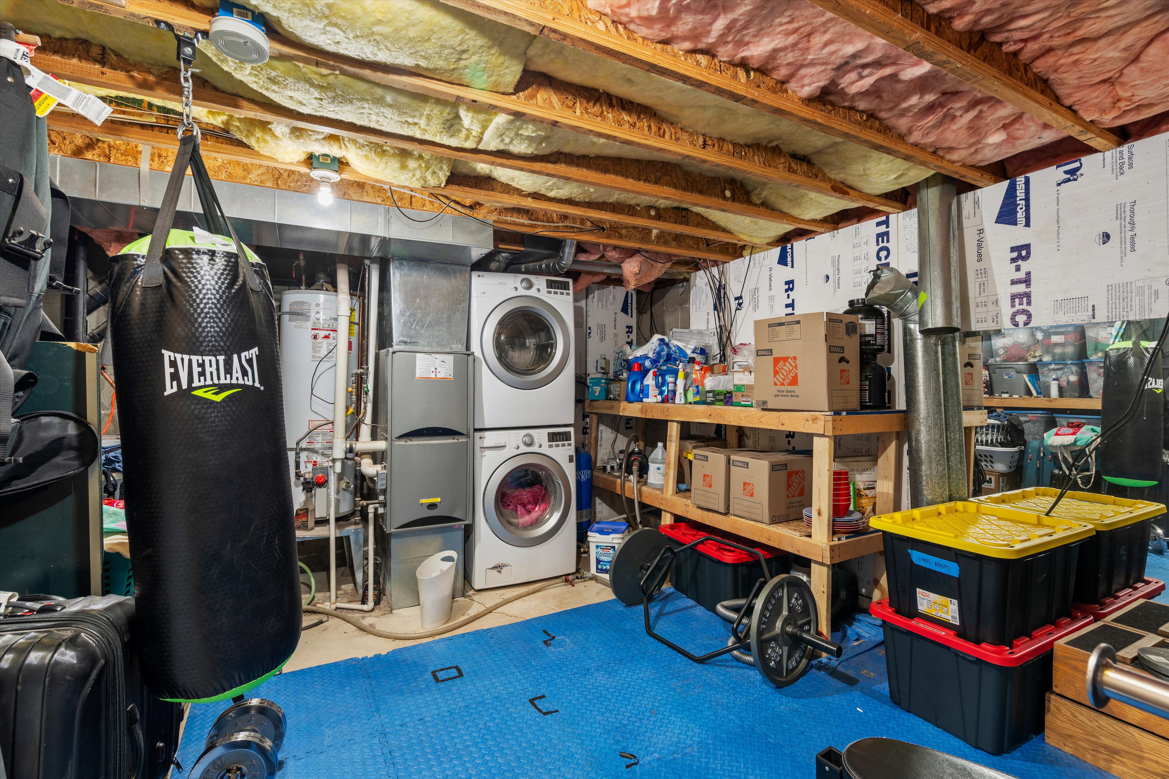 Unfinished basement space featuring exposed ceiling joists with insulation, a stacked washer and dryer, a water heater, and blue floor mats