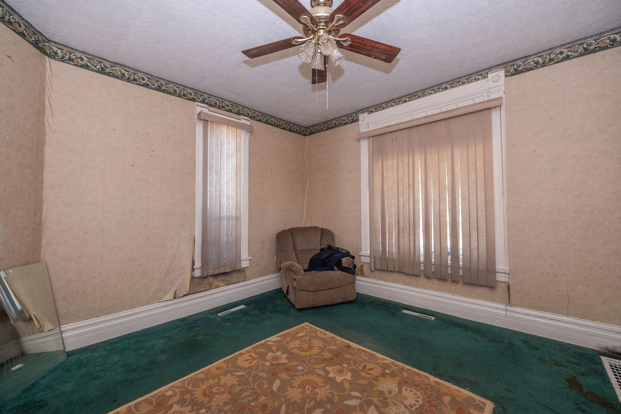 Room featuring two windows with vertical blinds, a ceiling fan with light fixture, decorative wallpaper border, and green carpet flooring