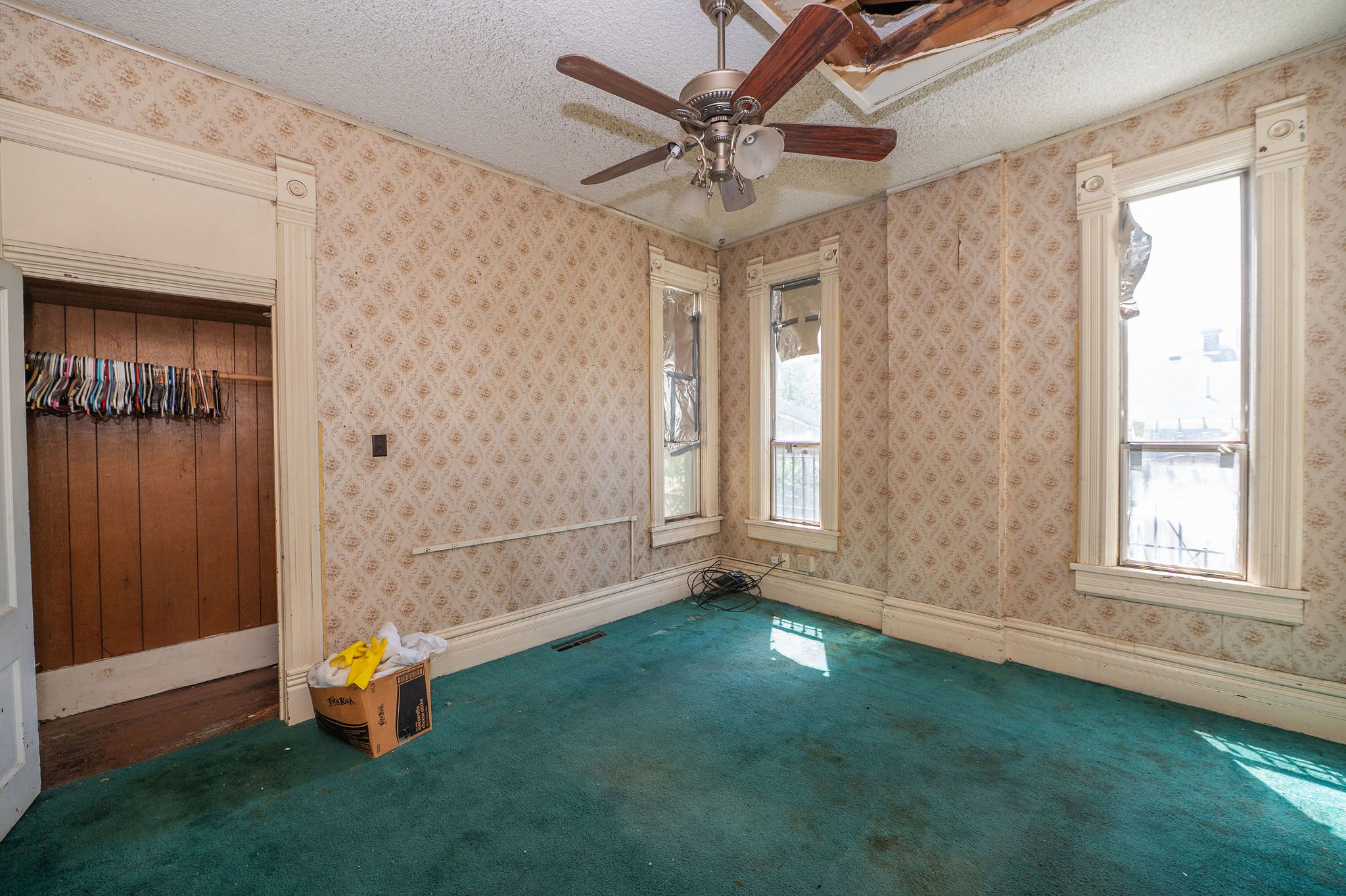 Room featuring patterned wallpaper, significant window trim, an open closet with wood paneling, a ceiling fan, and teal carpeting