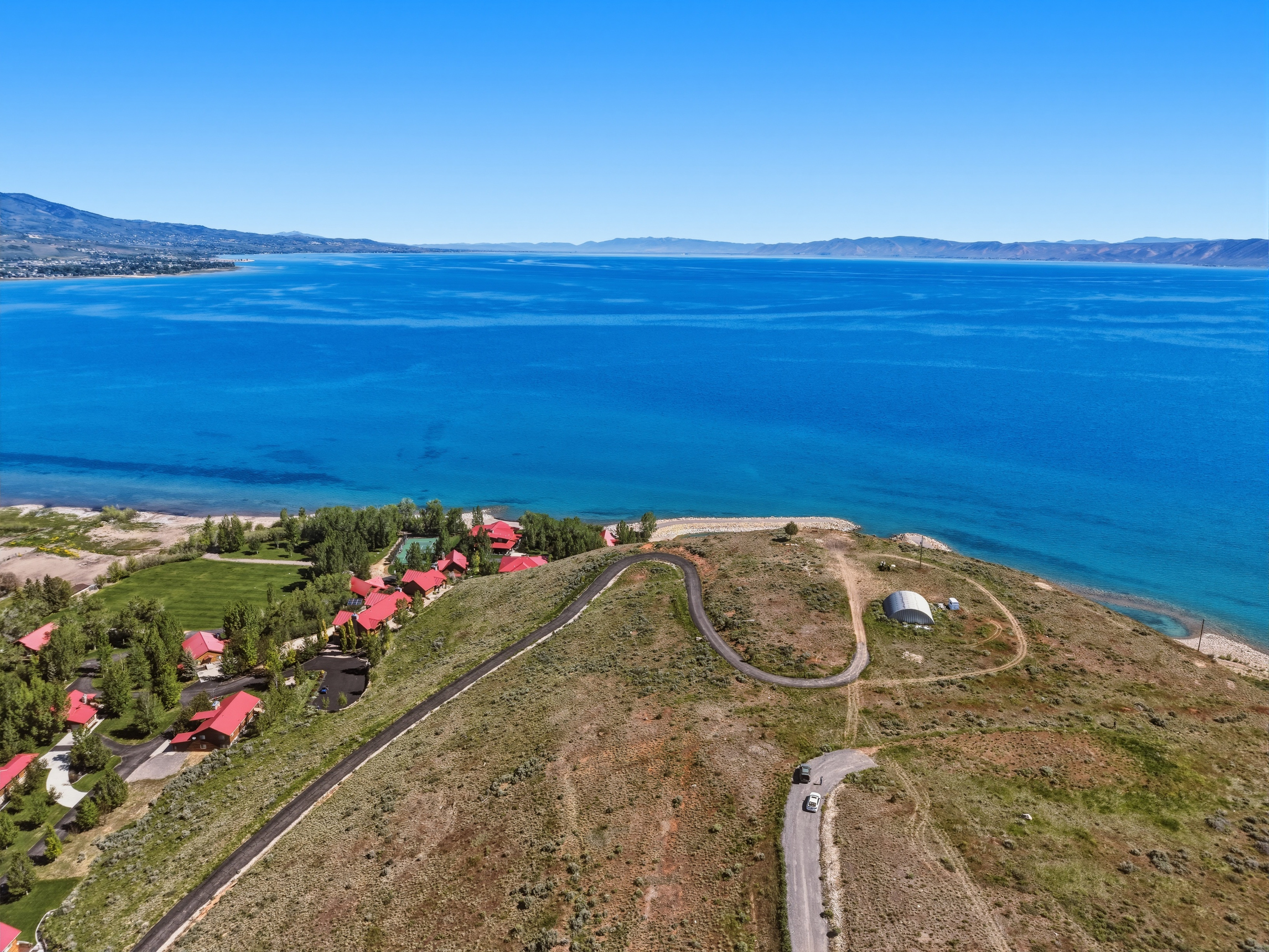 Expansive lakefront property featuring a winding paved road, lush green areas, and multiple structures with red roofs