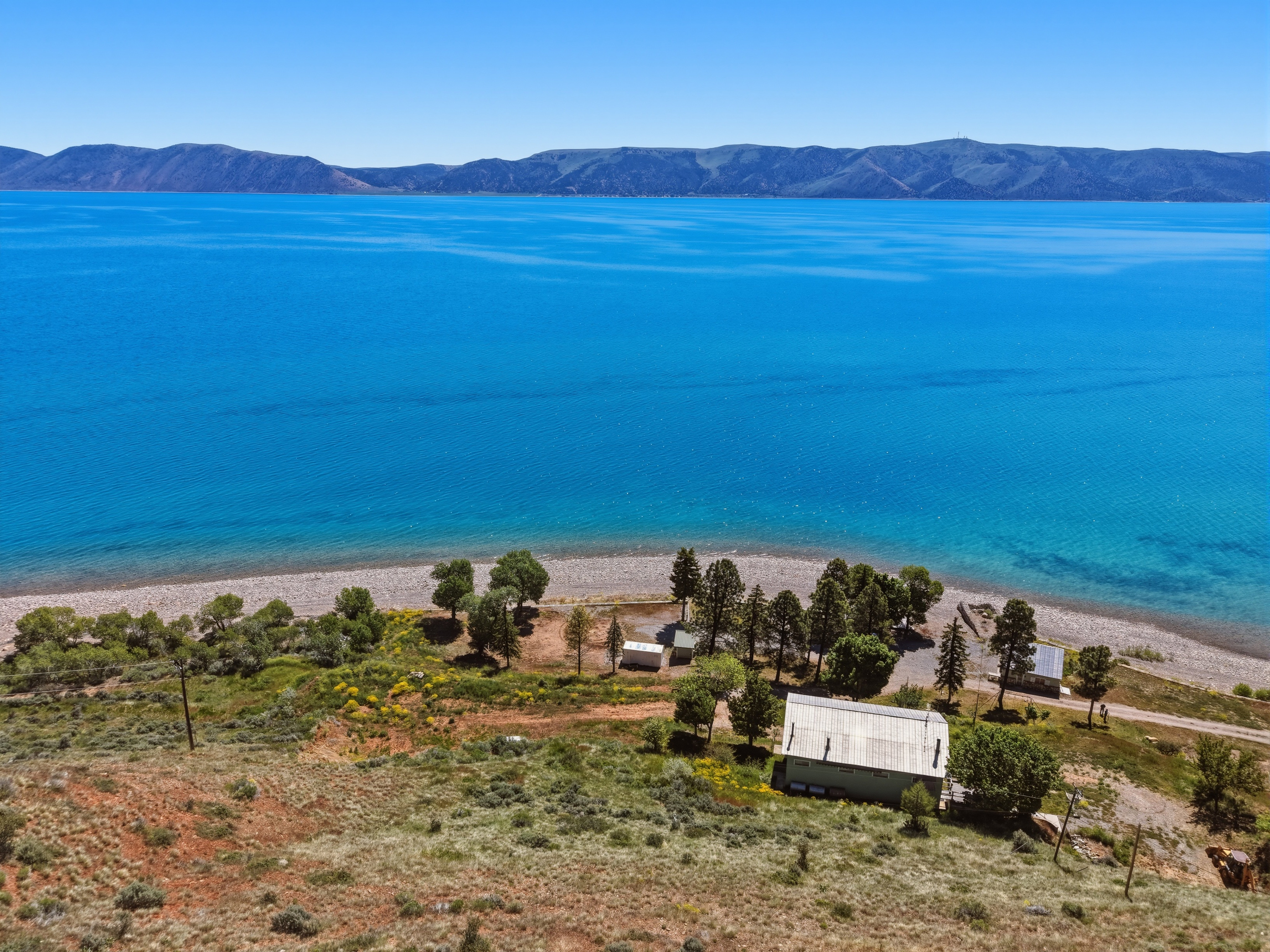 Waterfront property featuring a structure with a light-colored roof, surrounded by mature trees and natural landscaping, with a gravel beach at the water's edge and distant mountains
