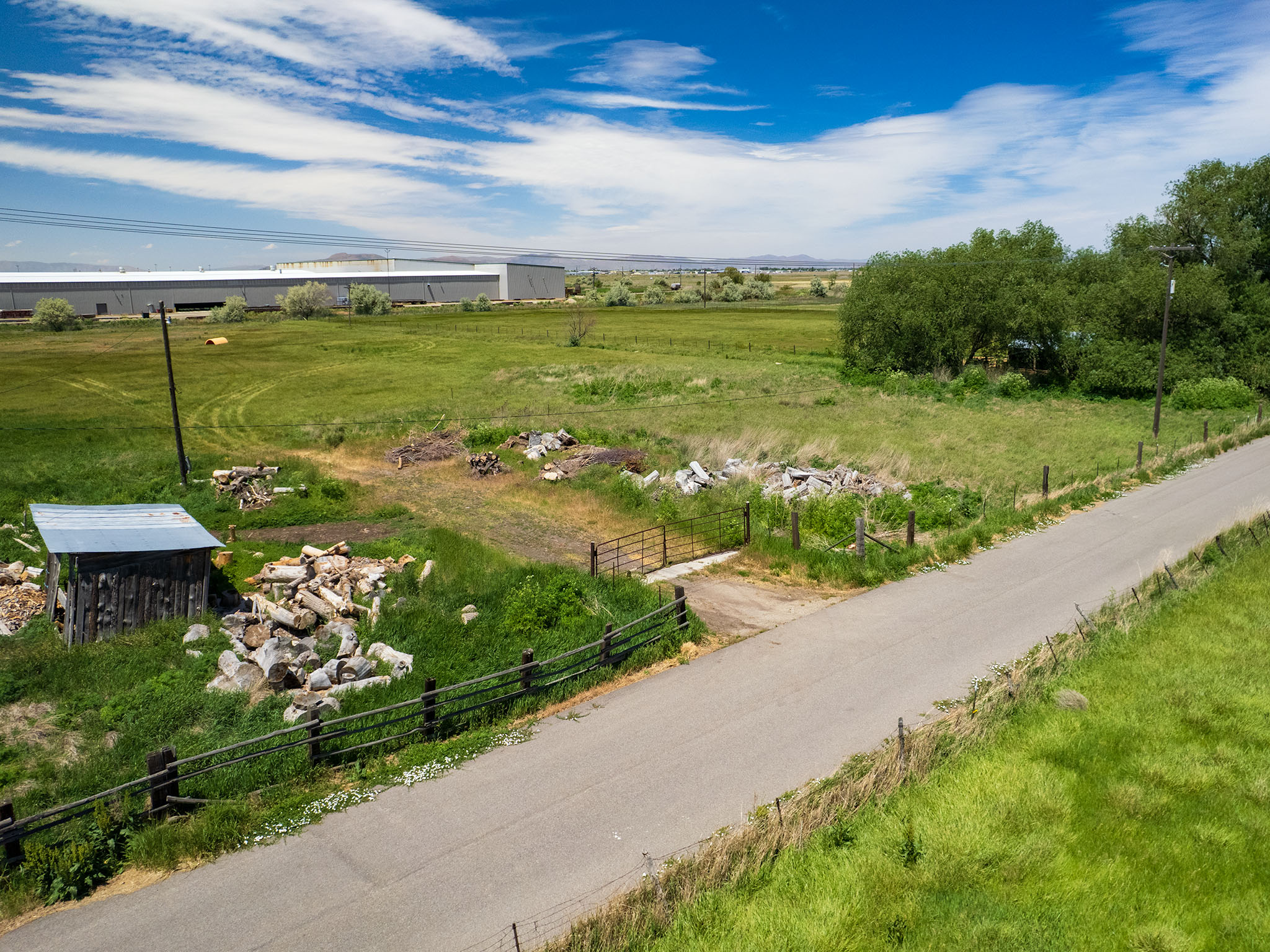 Expansive green acreage featuring a paved road, mature trees, and a distant industrial building under a bright blue sky