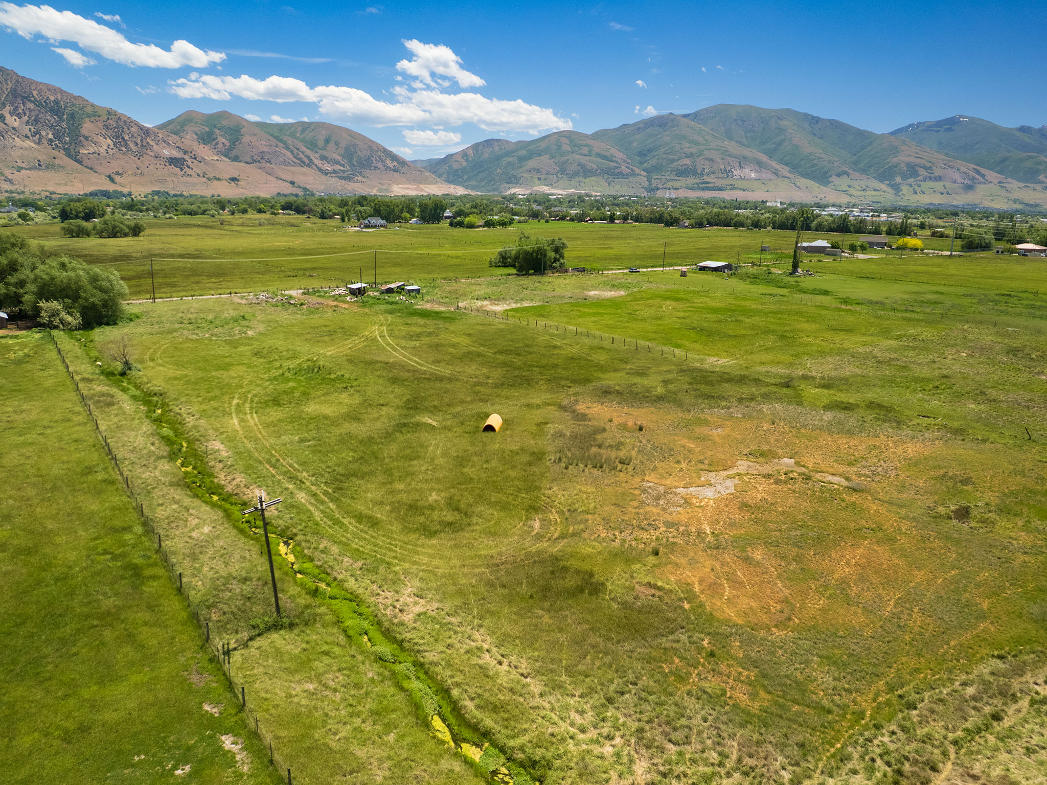 Expansive green fields featuring a clear mountain backdrop, a blue sky with scattered clouds, and scattered buildings in the distance
