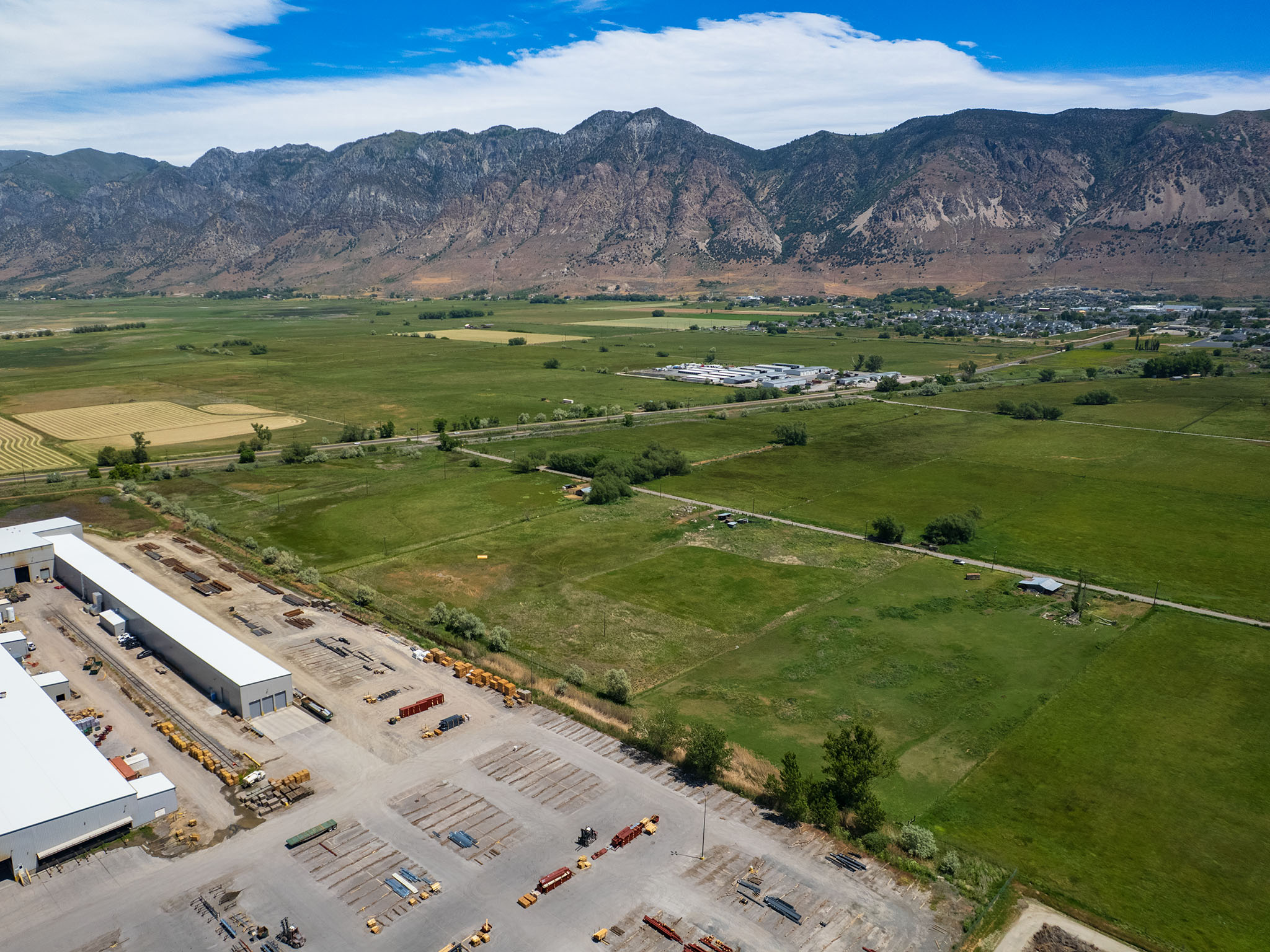 Expansive mountain range backdrop framing agricultural fields and a developed commercial property with multiple large warehouse structures, extensive paved parking, and visible storage areas