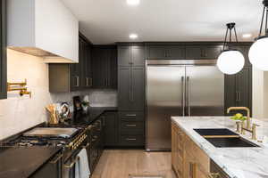 Gourmet kitchen featuring dark cabinetry, stainless steel appliances, black countertops, a pot filler, and a large central island with a white marble-patterned countertop and an undermount sink