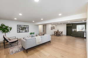 Open-concept living area featuring light wood-finish flooring, recessed lighting, white walls, a dining area, and a kitchen with dark green cabinetry and a window above the sink