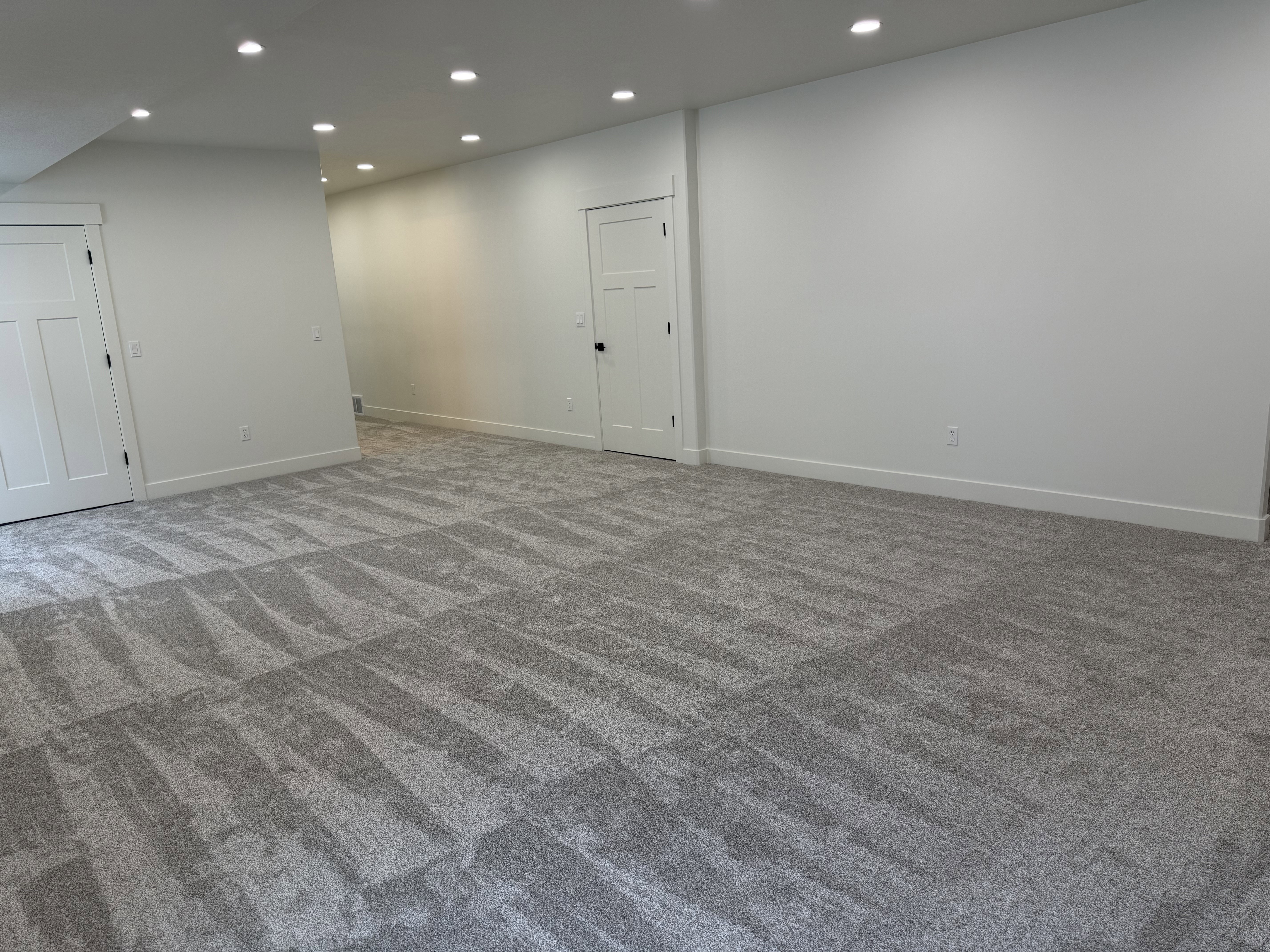 Spacious room featuring gray patterned carpet, recessed ceiling lighting, crisp white walls, and two white panel doors with black hardware