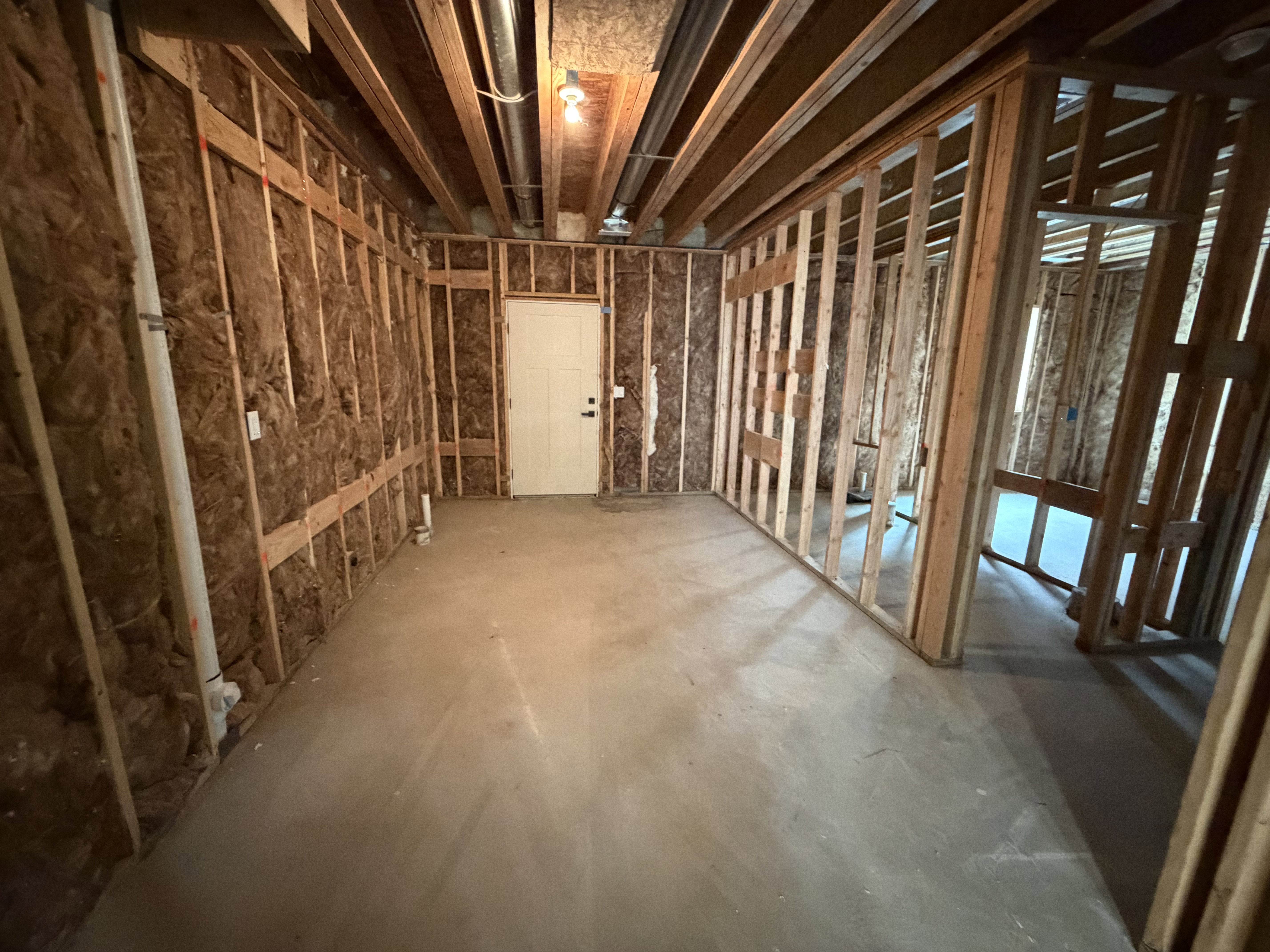 Unfinished basement space featuring exposed wood framing, concrete flooring, visible insulation, and a white interior door