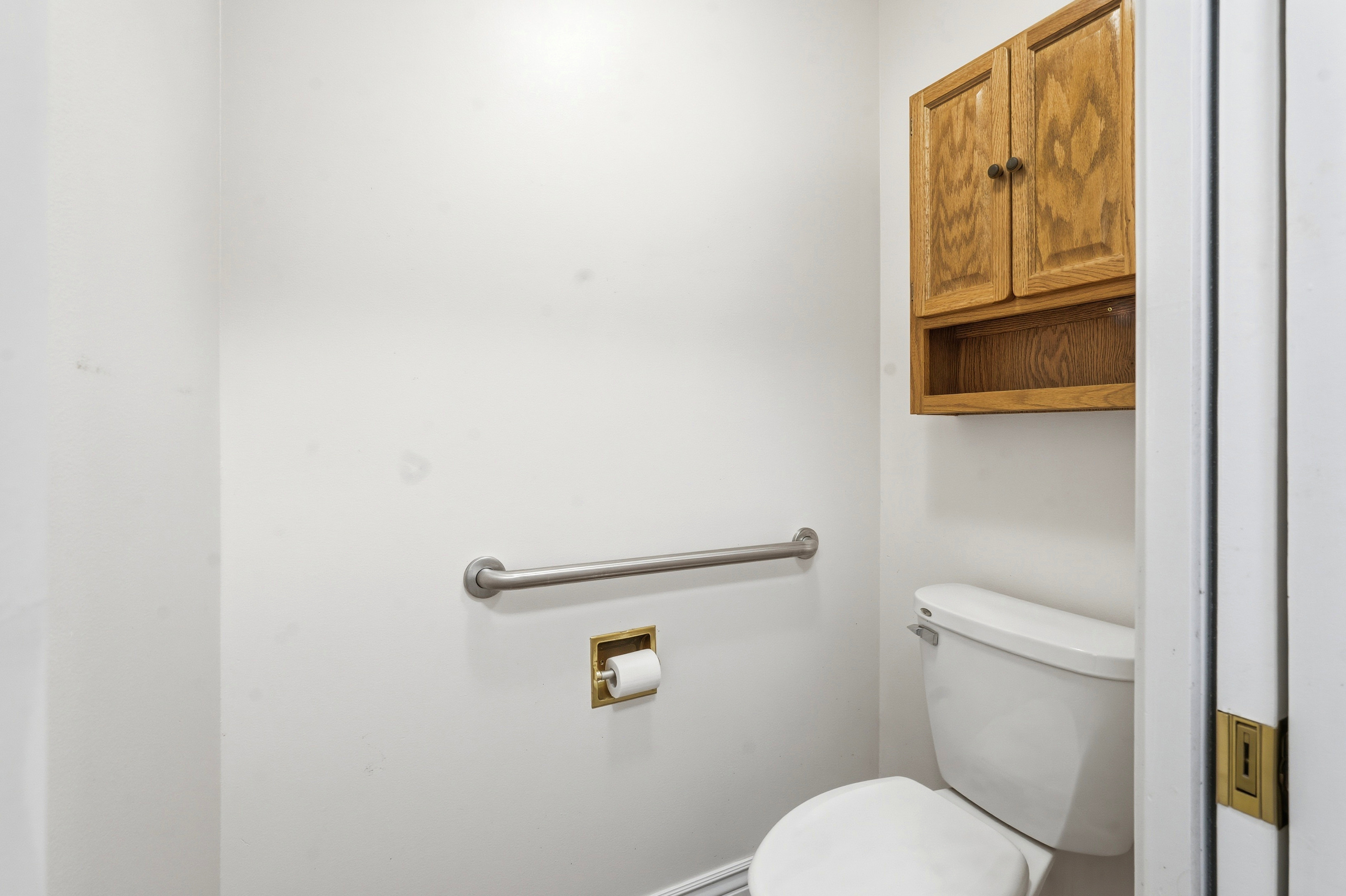 White toilet with wall-mounted wooden cabinetry, a brass-finish toilet paper holder, and a metal grab bar