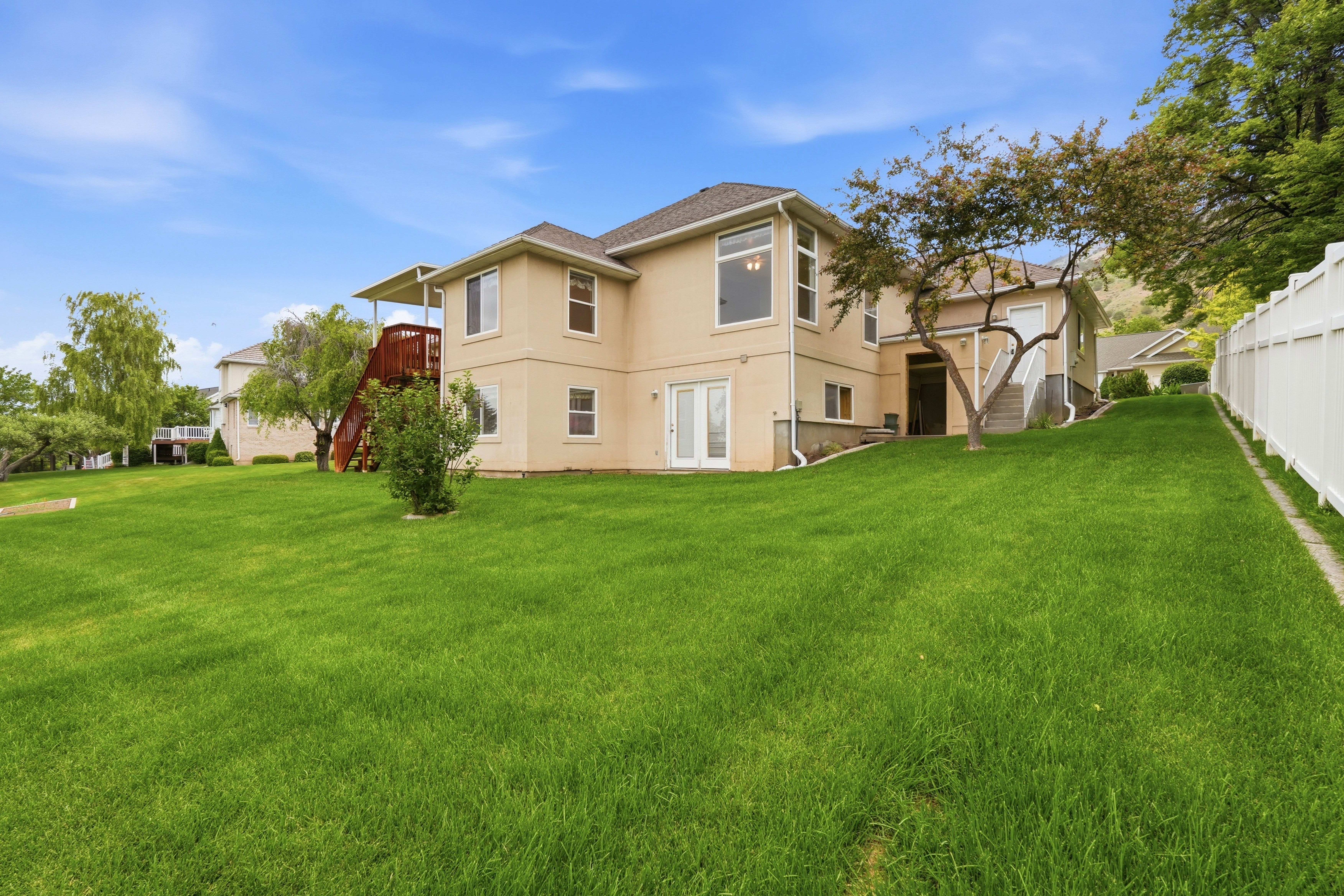 Expansive rear yard featuring a manicured lawn, stucco exterior, multiple windows, double French doors, and an upper-level deck with stairs