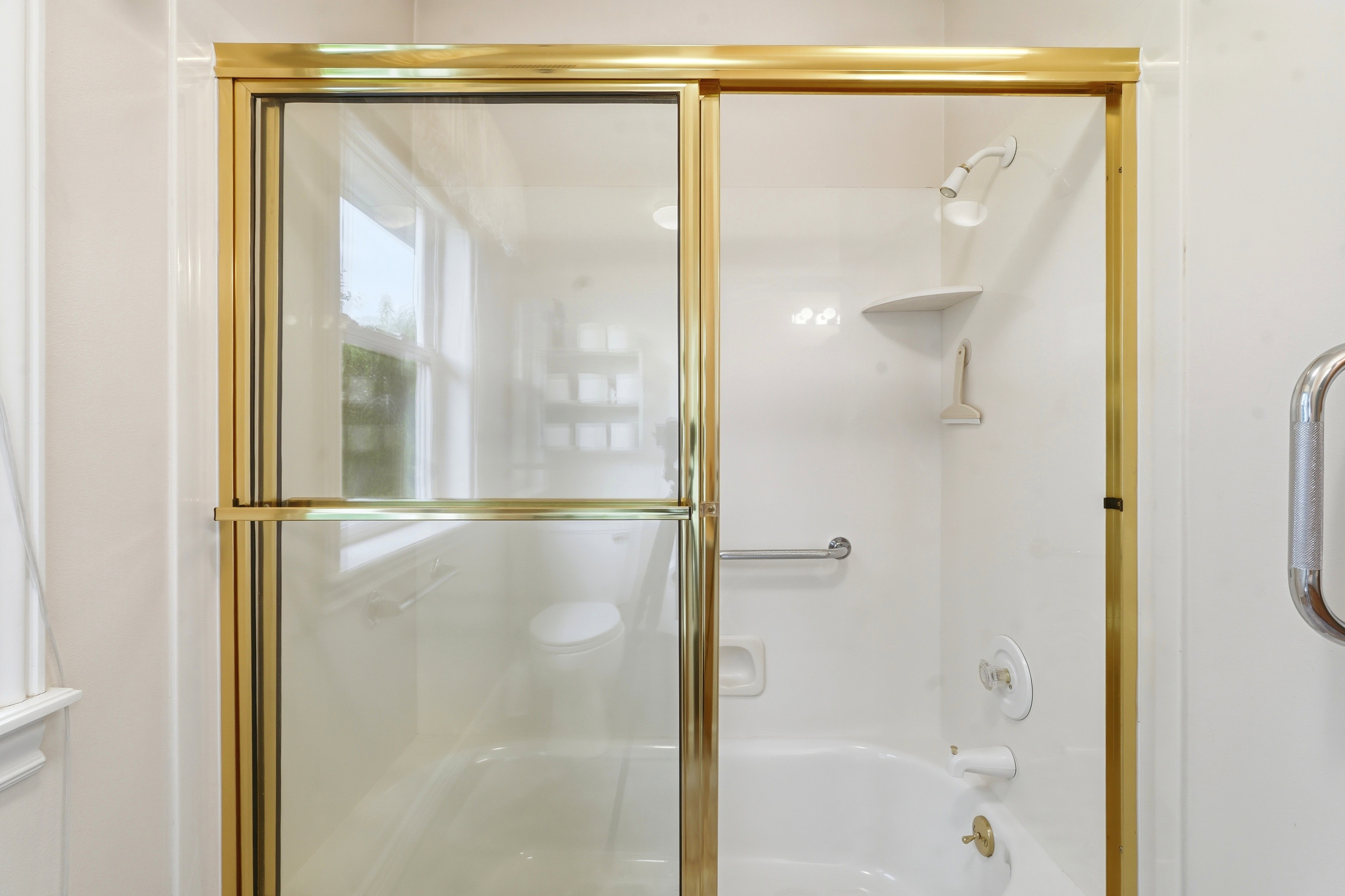 Bathtub and shower combination featuring a gold-framed glass enclosure, white acrylic surround, integrated corner shelf, and grab bar